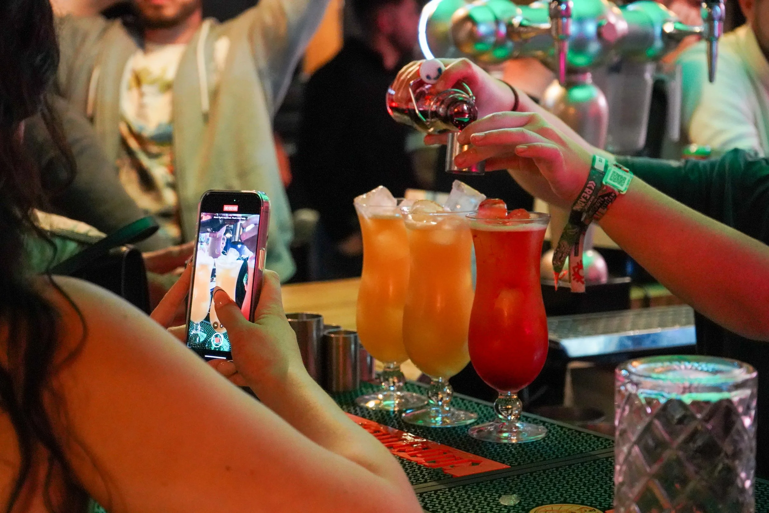 A person captures a photo of colorful cocktails on a bar with their smartphone as another person pours a drink, with other bar patrons in the background.