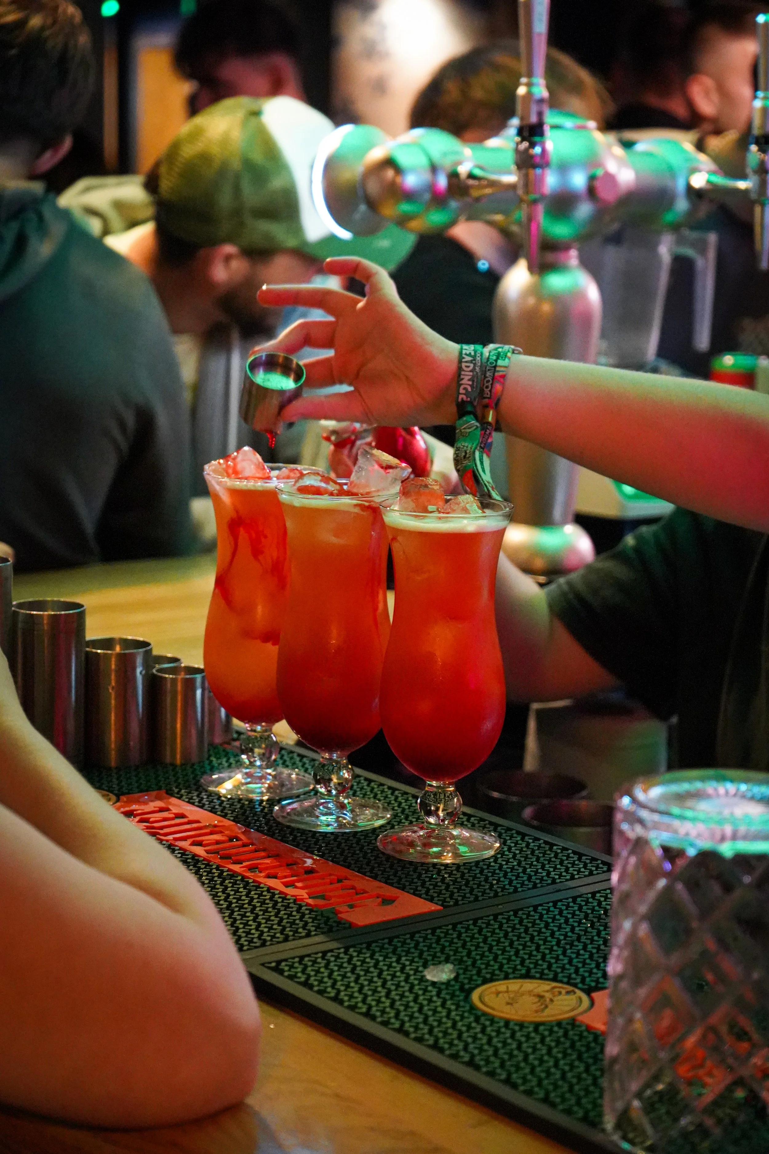 A bartender pouring a red drink into Hurricane cocktail glasses on a bar counter with several drinks already served. People are gathered in the background behind the bar.