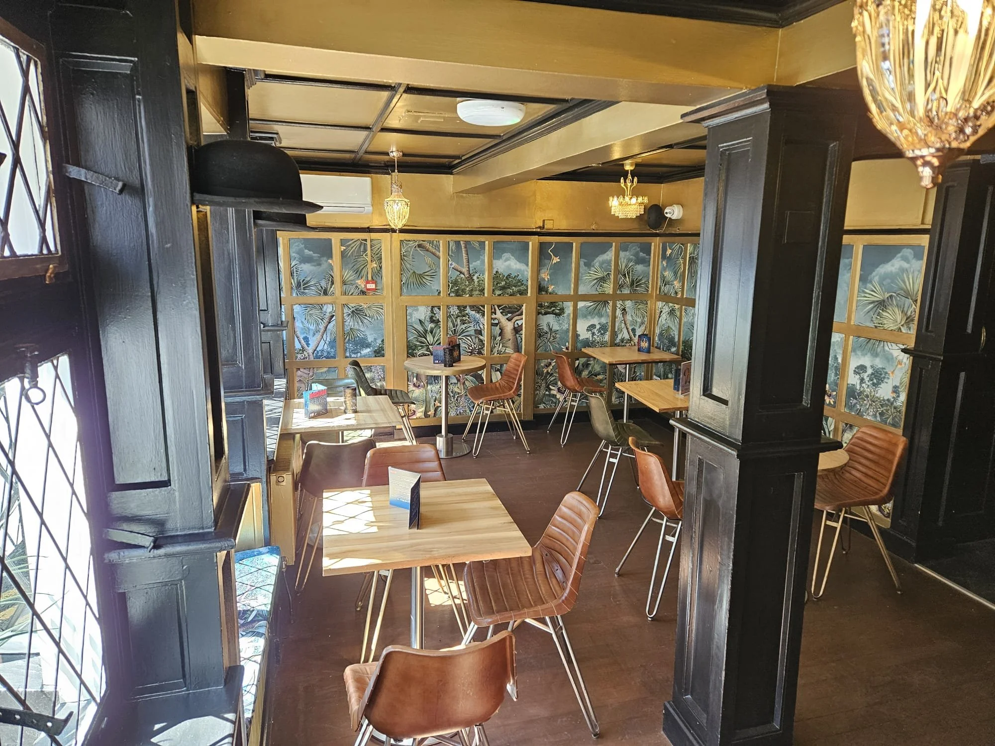 Interior of a restaurant with wooden tables and chairs, black wood paneling, and a scenic wall mural of a tropical landscape with palm trees.