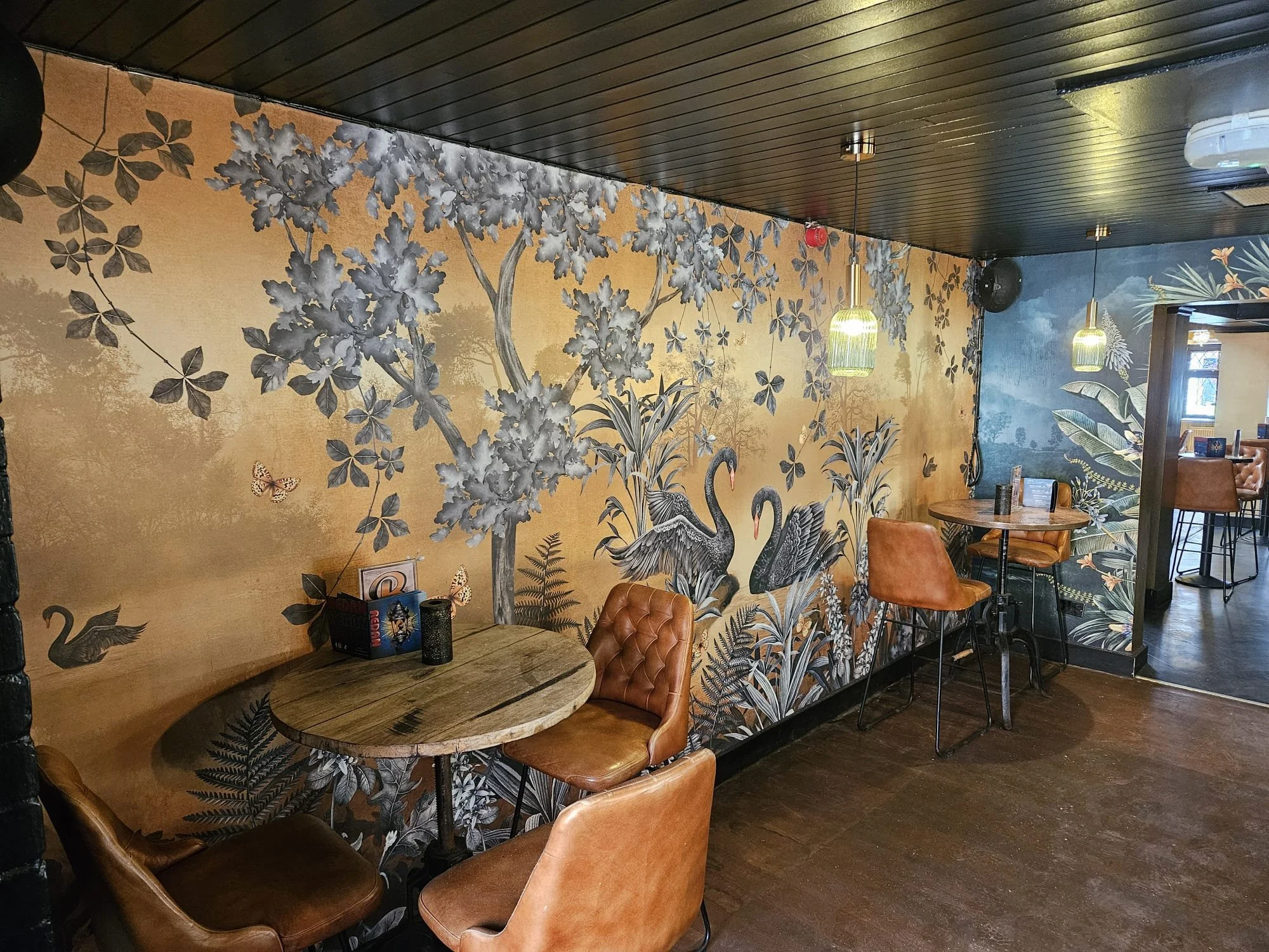 Cozy restaurant interior with a nature-themed mural featuring trees, leaves, ferns, and swans on a gold background, illuminated by pendant lights, with seating including leather chairs and wooden tables.
