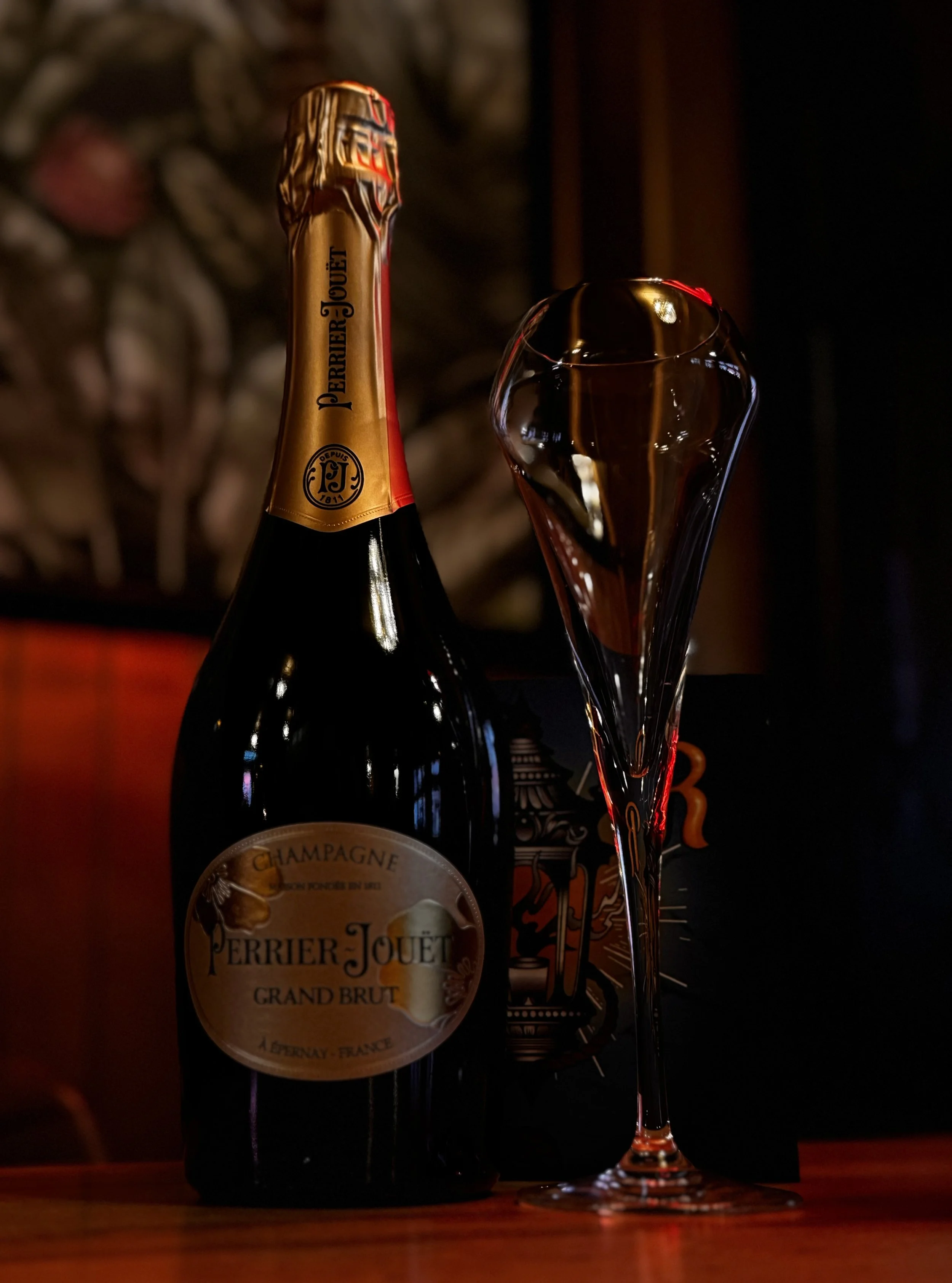 A bottle of Perrier-Jouët Grand Brut champagne with a champagne flute filled with champagne next to it, on a wooden surface with a dark background.
