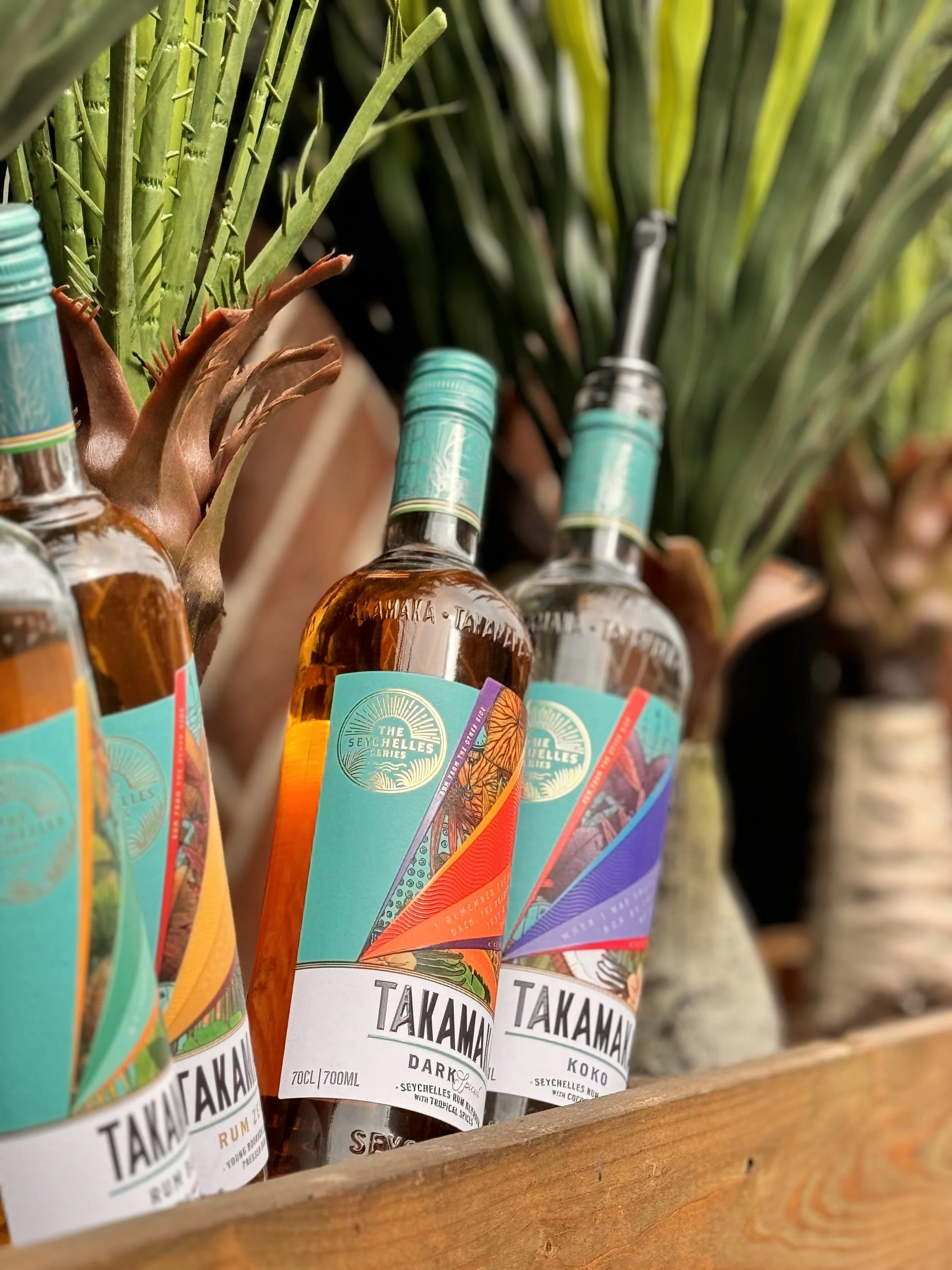 Three bottles of Takamaka rum labeled with tropical designs, placed on a wooden surface among palm trees.