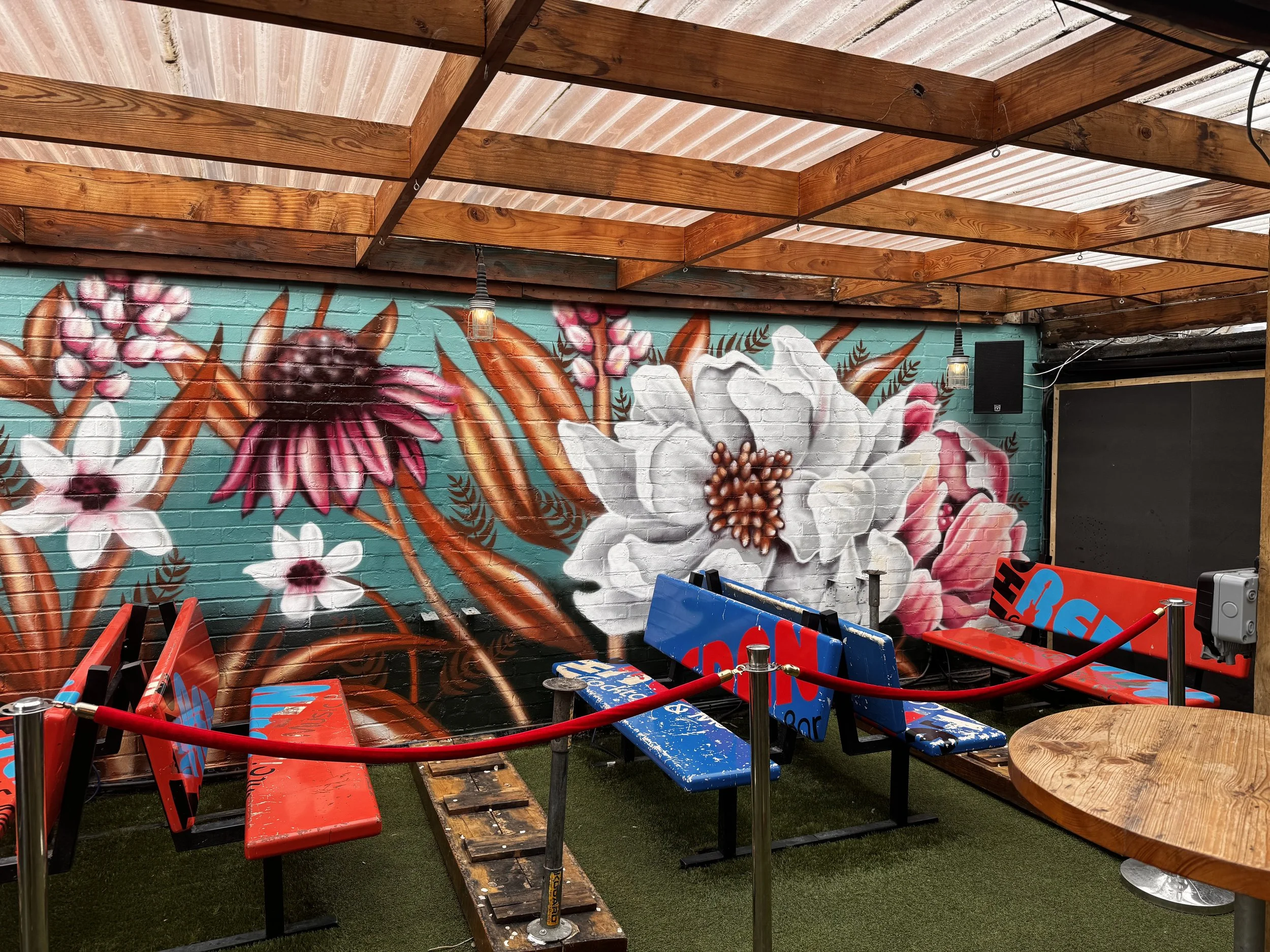 floral wall mural, featuring large white, pink, and purple flowers, against a turquoise brick background. Surrounded by red velvet ropes, 3 heated benches and a round wooden table on the right. The space has a wooden roof and hanging light bulbs.