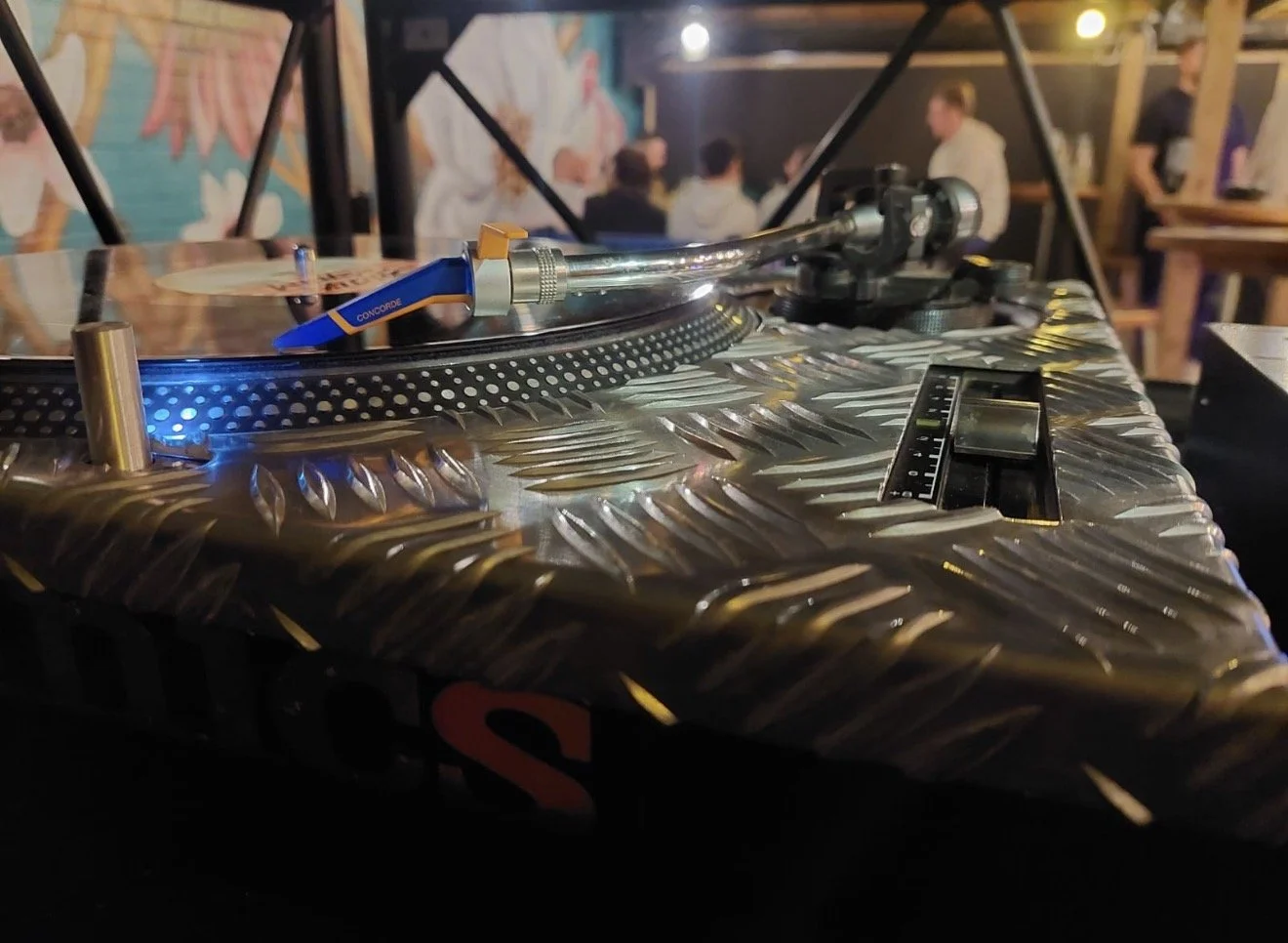 Close-up of a DJ turntable with a metallic surface, a tonearm, and a vinyl record, in a dimly lit setting with people in the background. Dj events every week with all medium of turntable, digital and vinyl.