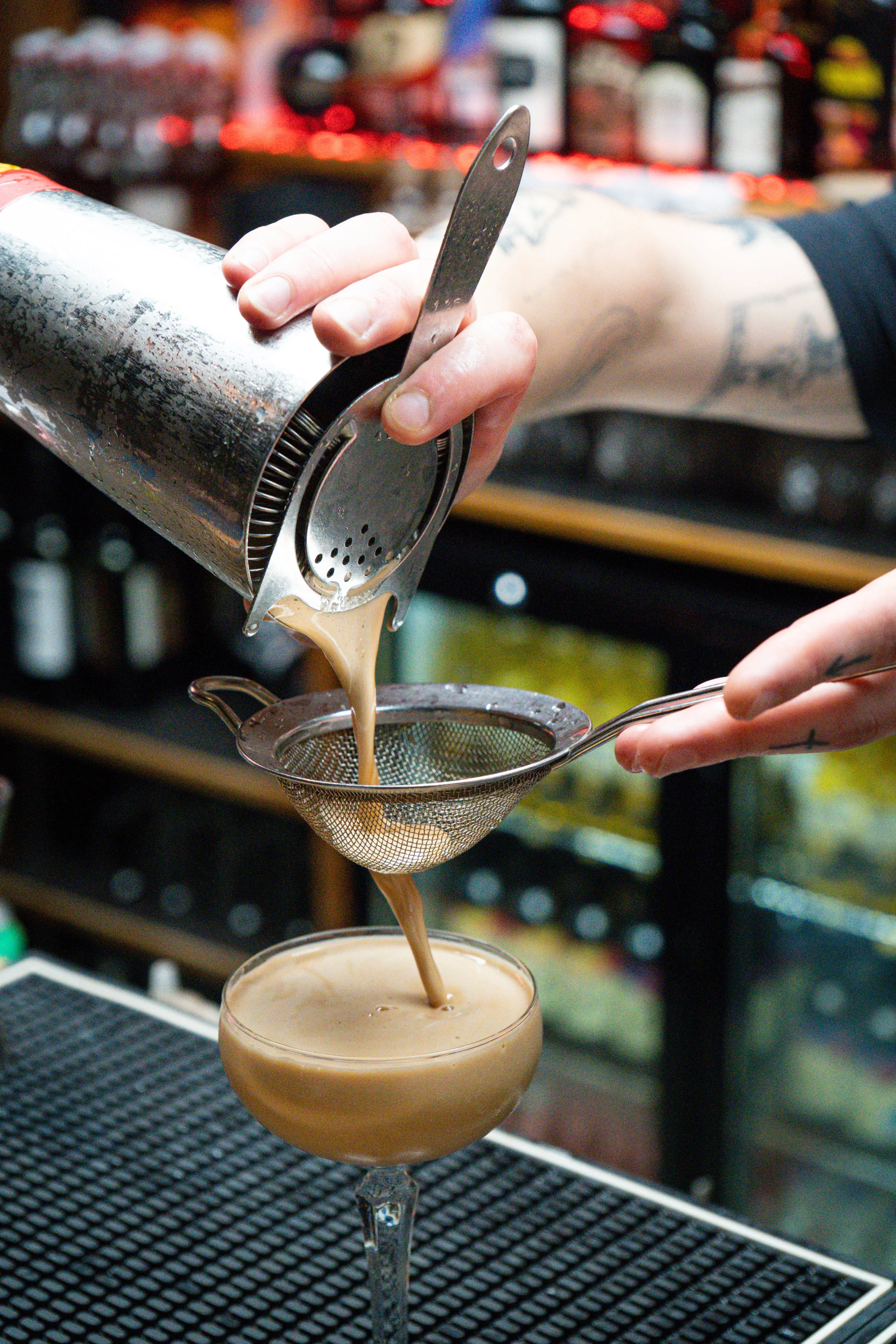 A bartender pours a creamy cocktail through a strainer into a coupe glass in a bar.