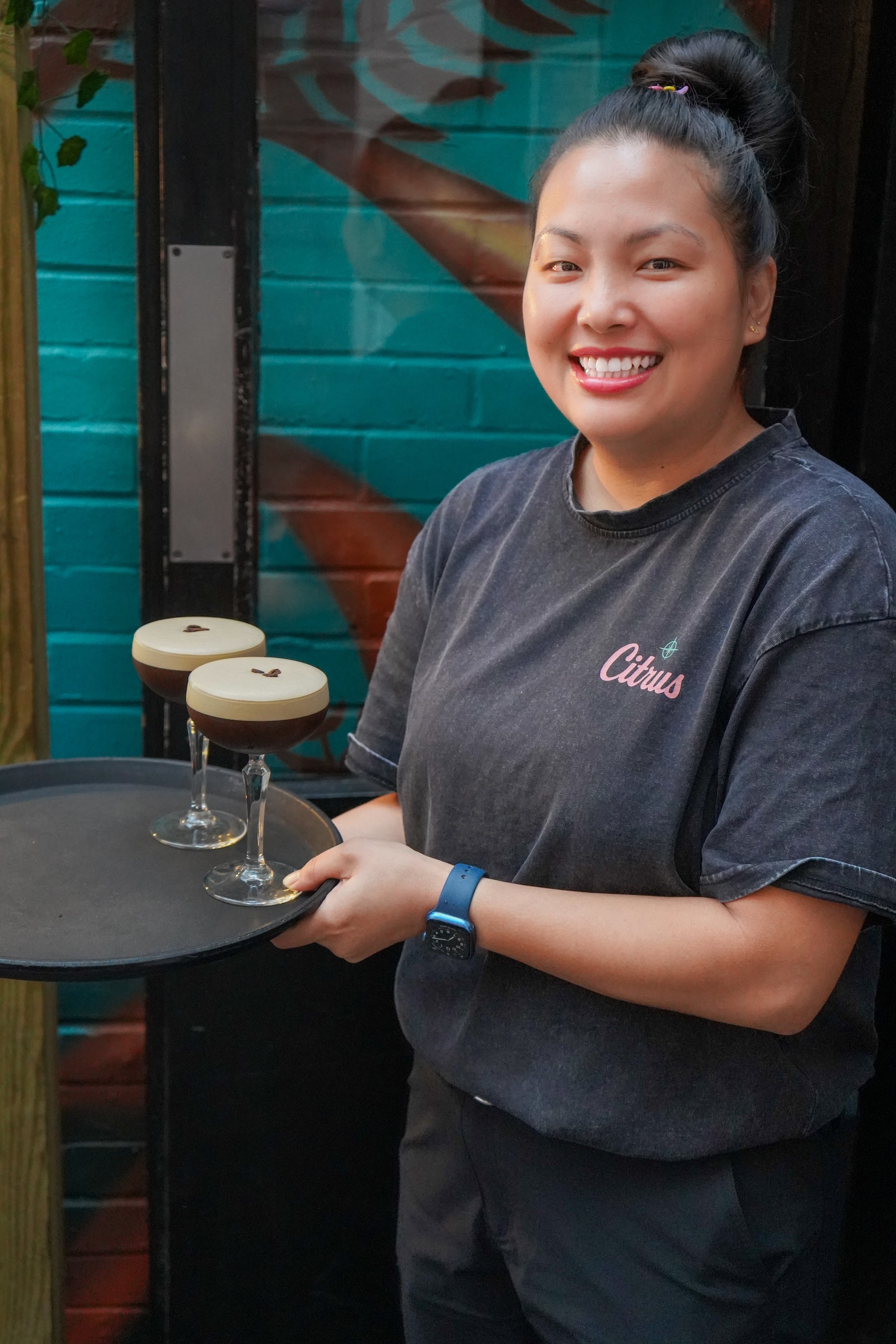A smiling woman in a dark gray T-shirt with "Citrus" written on it, holding a tray with two coffee drinks topped with whipped cream.