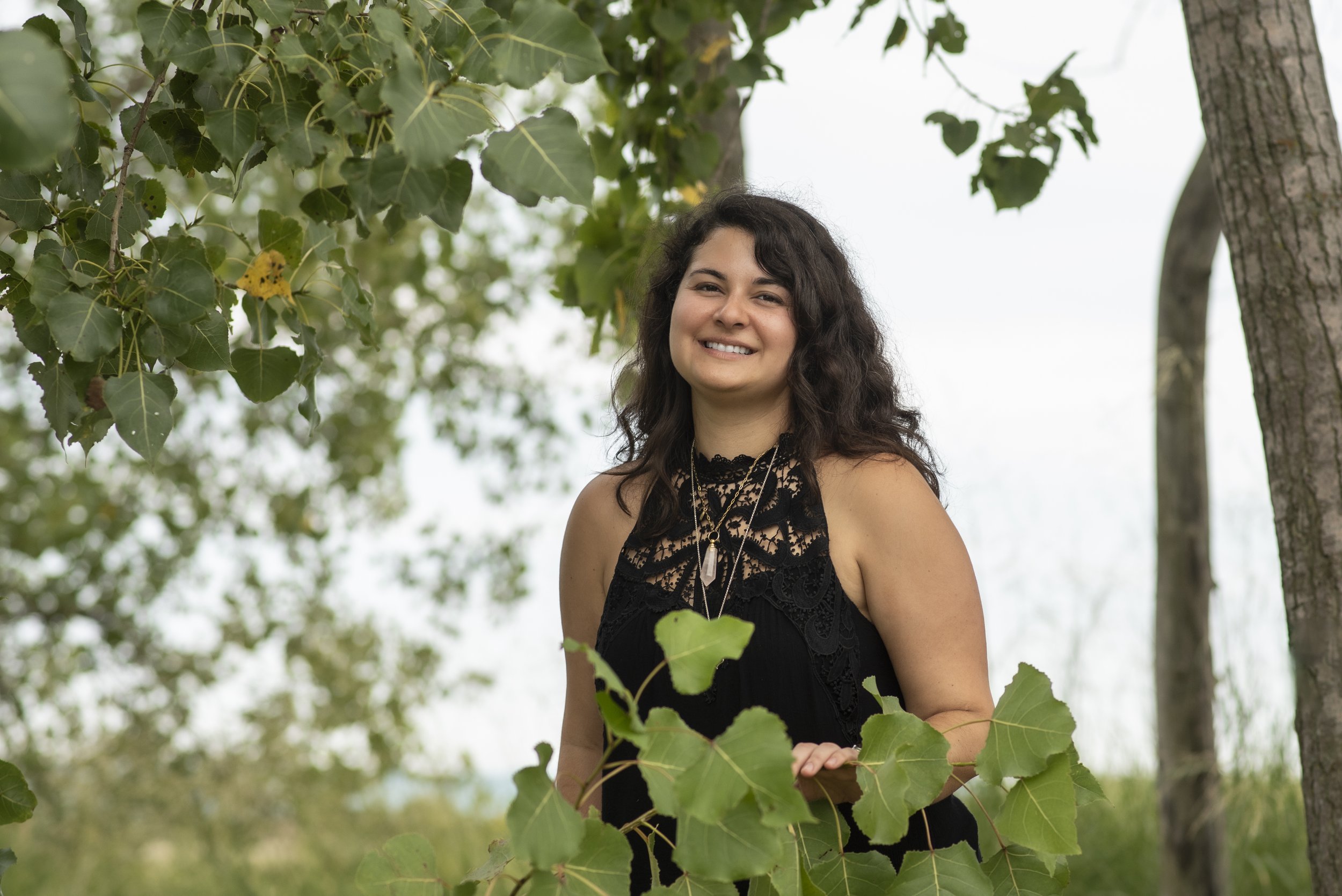 Portrait of Alexandra smiling and standing behind a bush as she holds a branch back to view the camera.