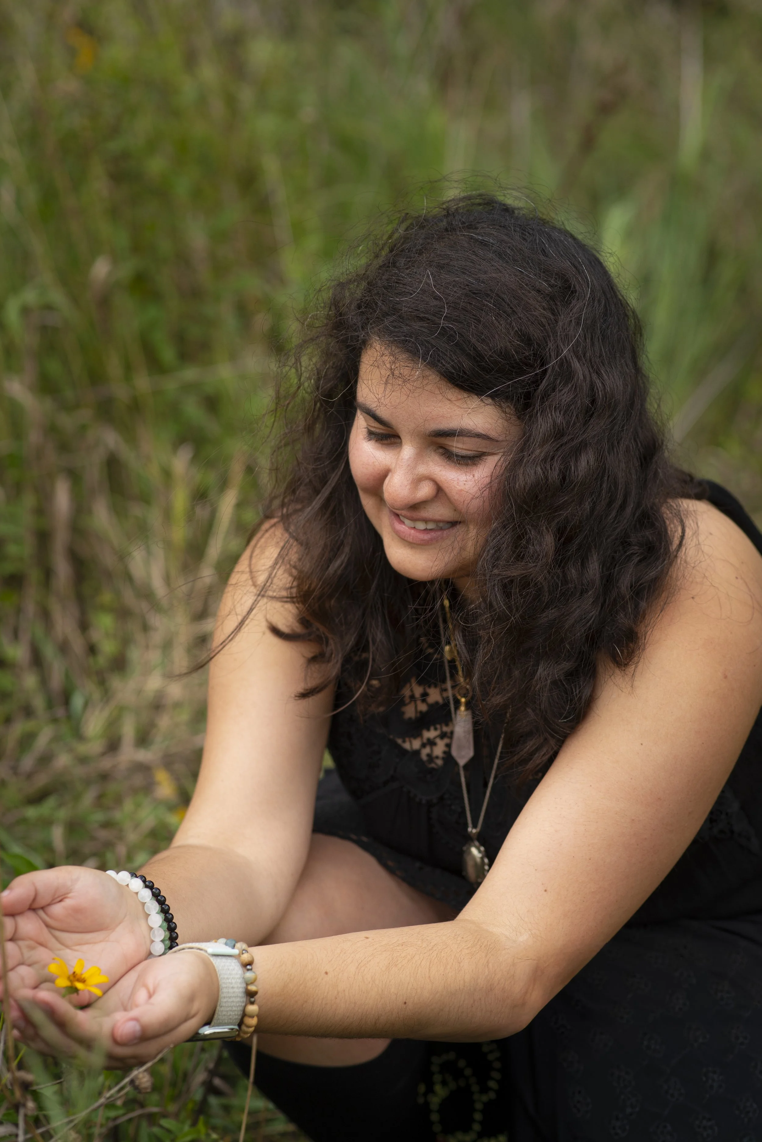 Alexandra kneeling in a field, smiling, while holding a flower as she performs reiki on it