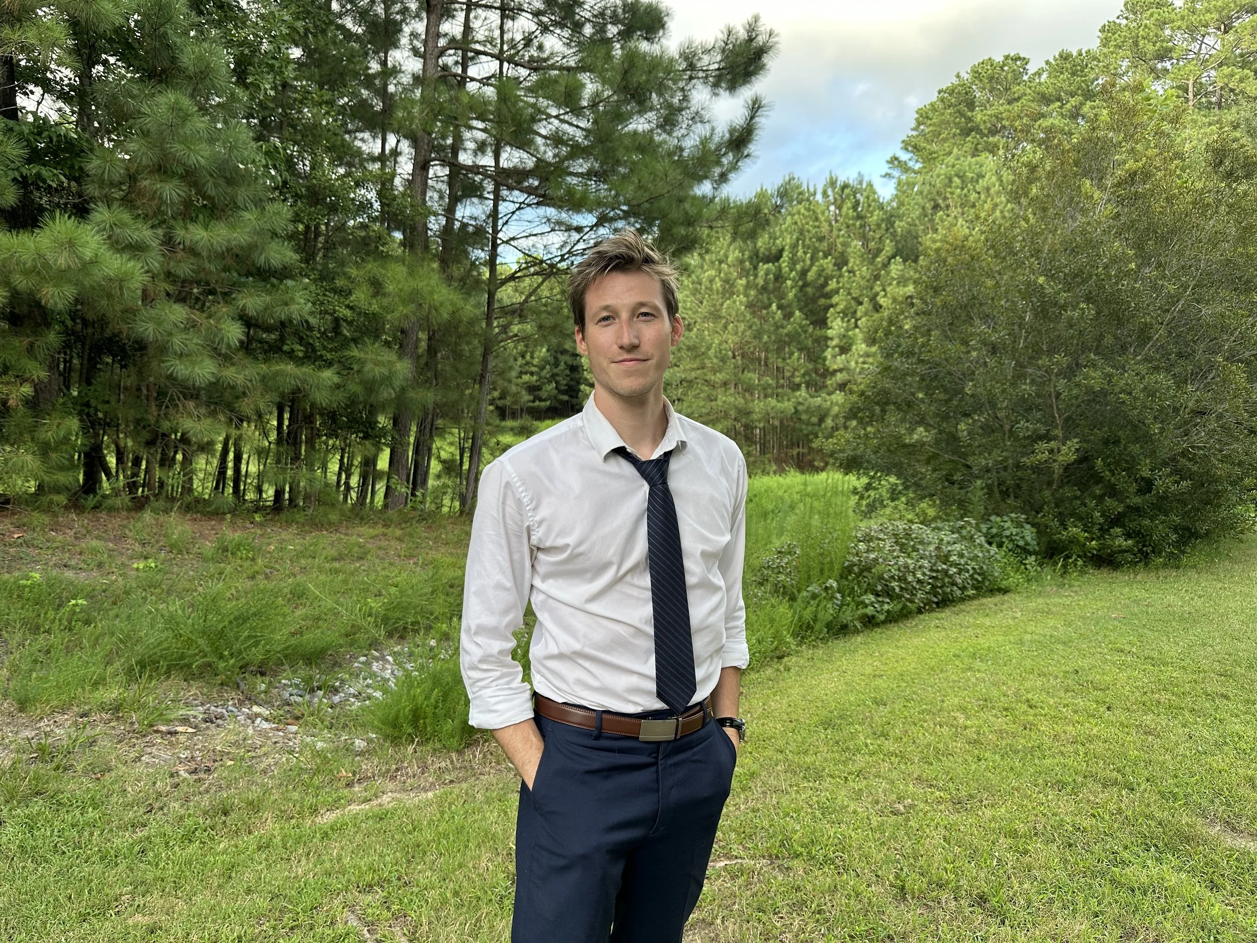 A young man in business attire standing outdoors in a grassy area surrounded by trees and greenery.