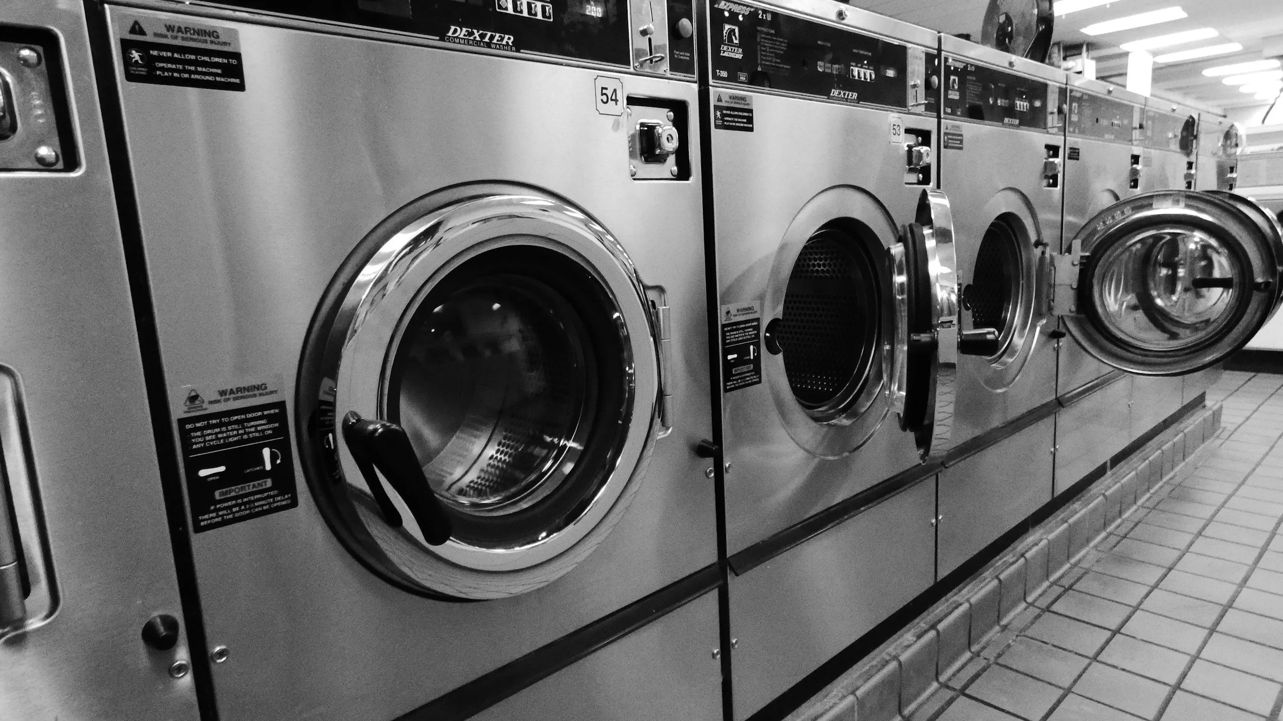 A row of silver commercial washing machines in a laundromat, with some doors open and others closed, on a tiled floor.