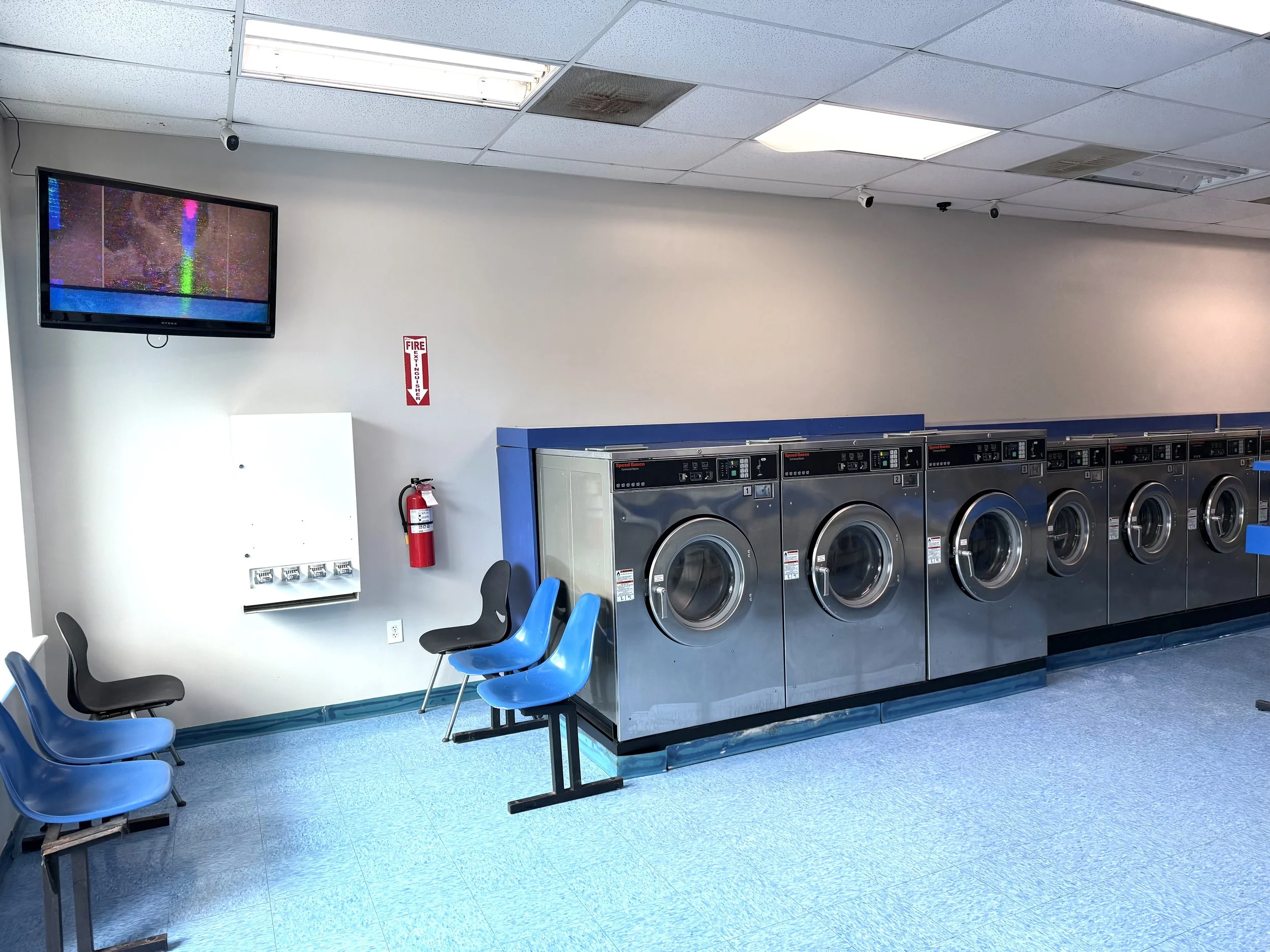 A laundromat with rows of stainless steel washing machines, a wall-mounted TV, chairs, a fire extinguisher, and a white utility box.