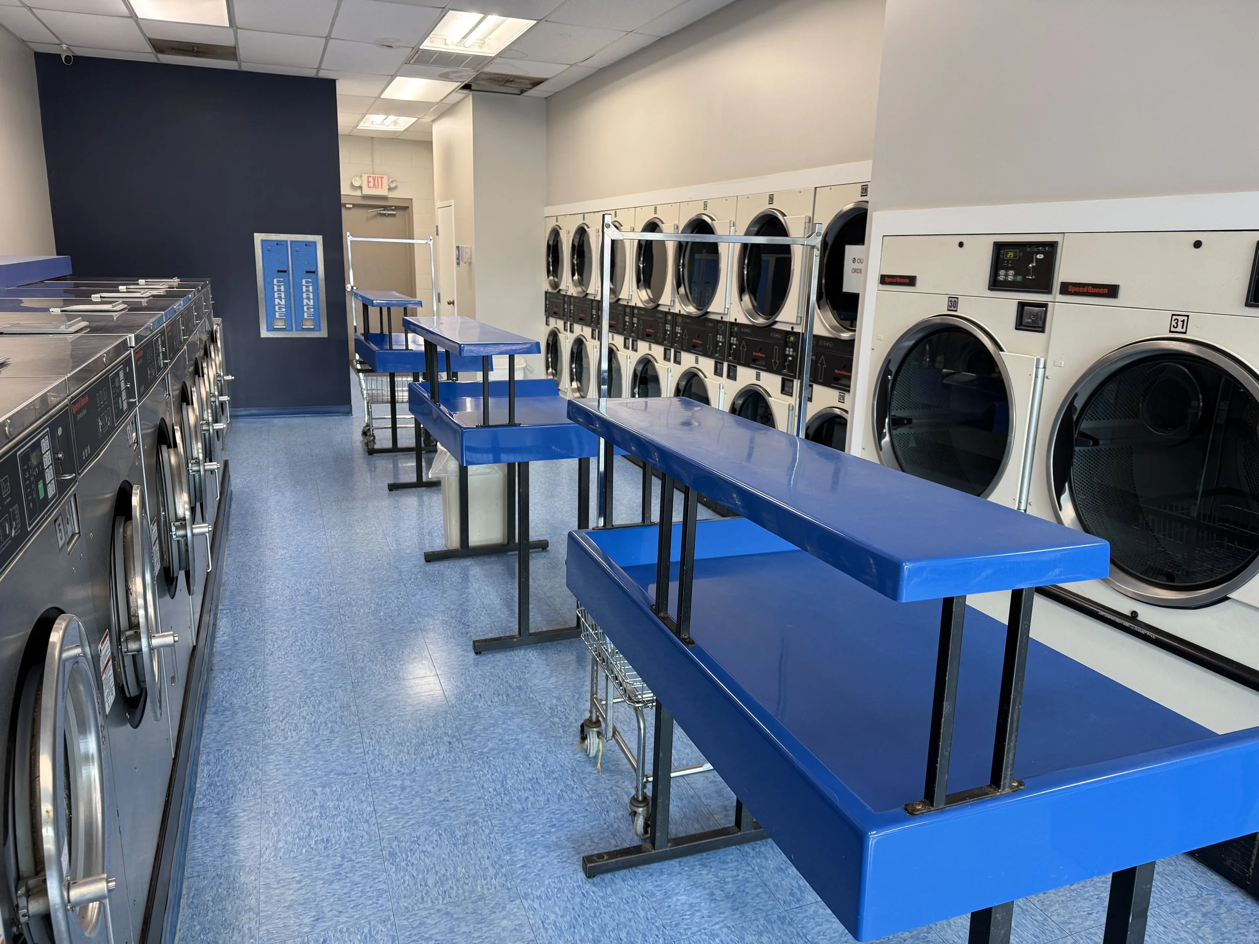Laundry room with several industrial washing machines and dryers along the walls, blue service carts in the center, and a blue tiled floor.
