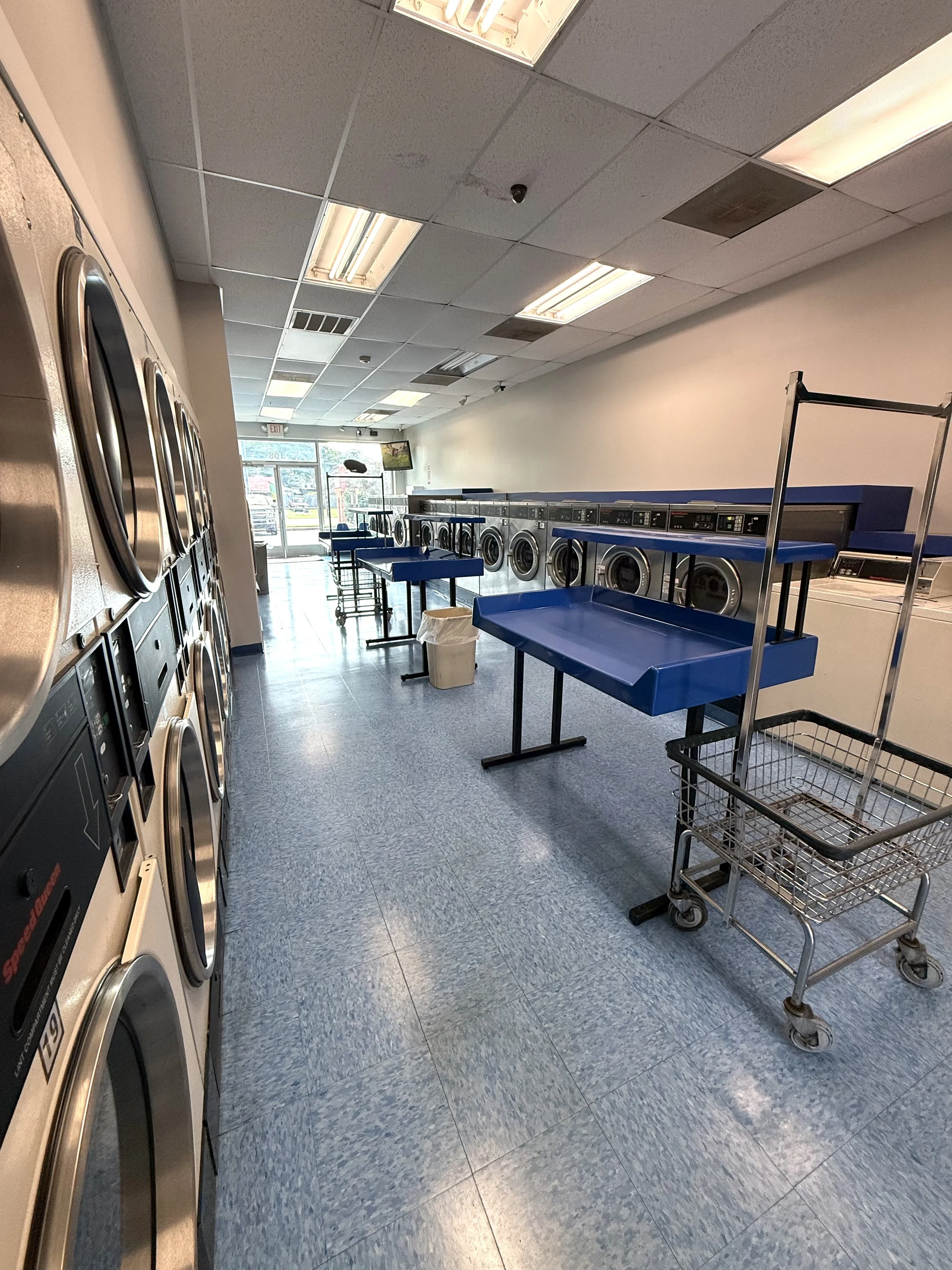 A laundromat with a row of front-loading washing machines on the right and dryers on the left, with blue folding tables and a shopping cart in the middle, near the door at the back.