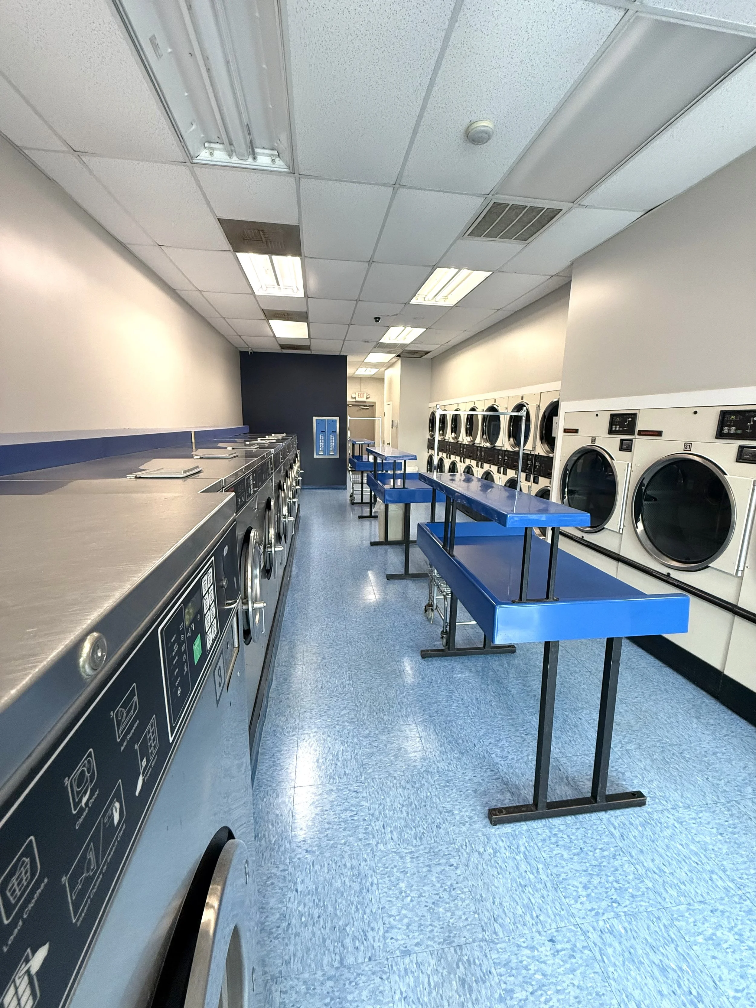 Empty laundromat with front-loading washing machines along the back wall and top-loading dryers on the left side, with blue folding tables in the middle.