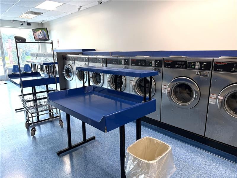 Self-service laundromat with washing machines, blue folding tables, laundry carts, a trash bin, and a TV mounted on the wall.