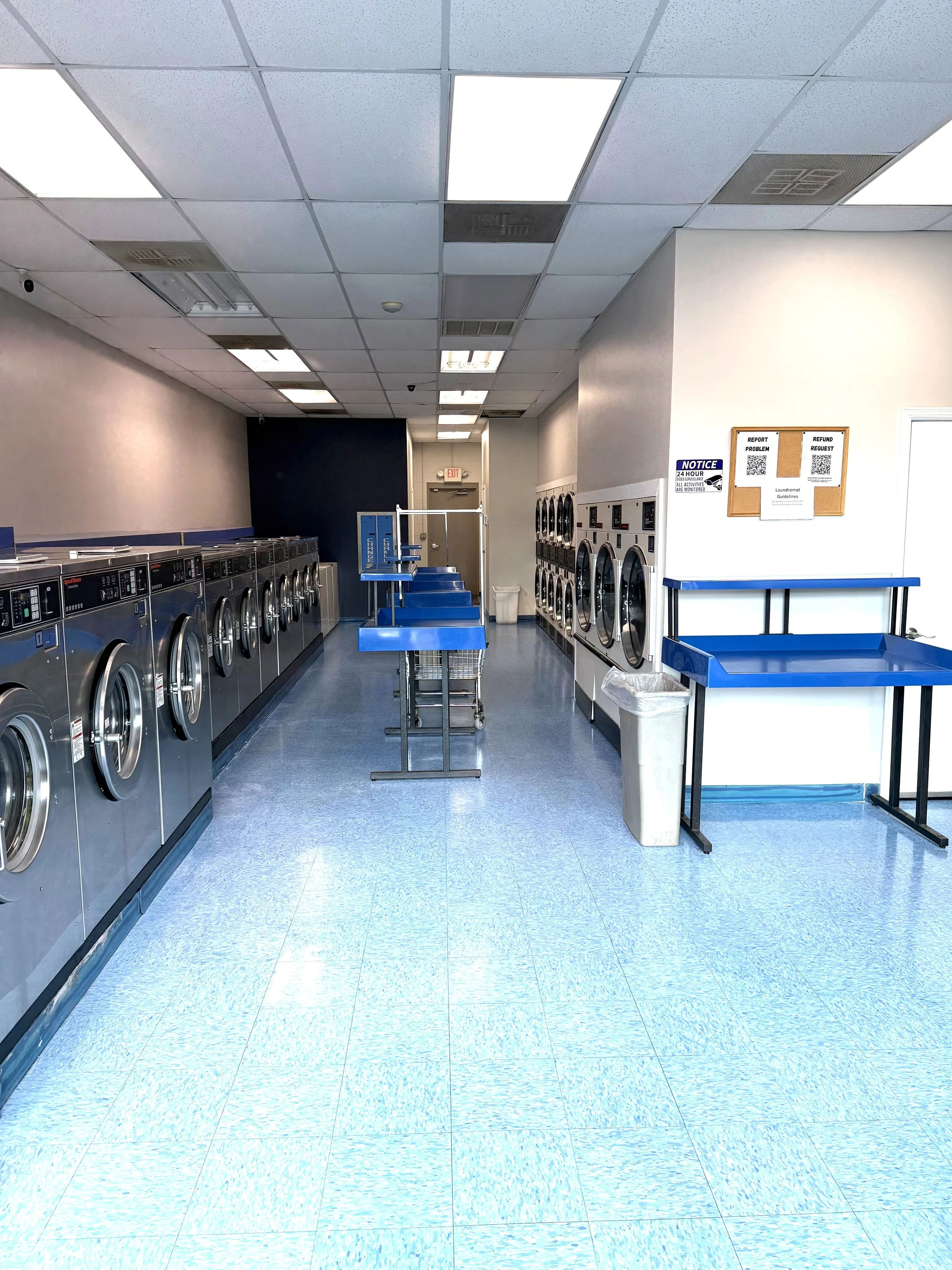 Empty laundromat with front-loading washers on the left and dryers on the right, blue folding tables and shelves in the middle, white walls, tiled blue flooring, and ceiling lights.