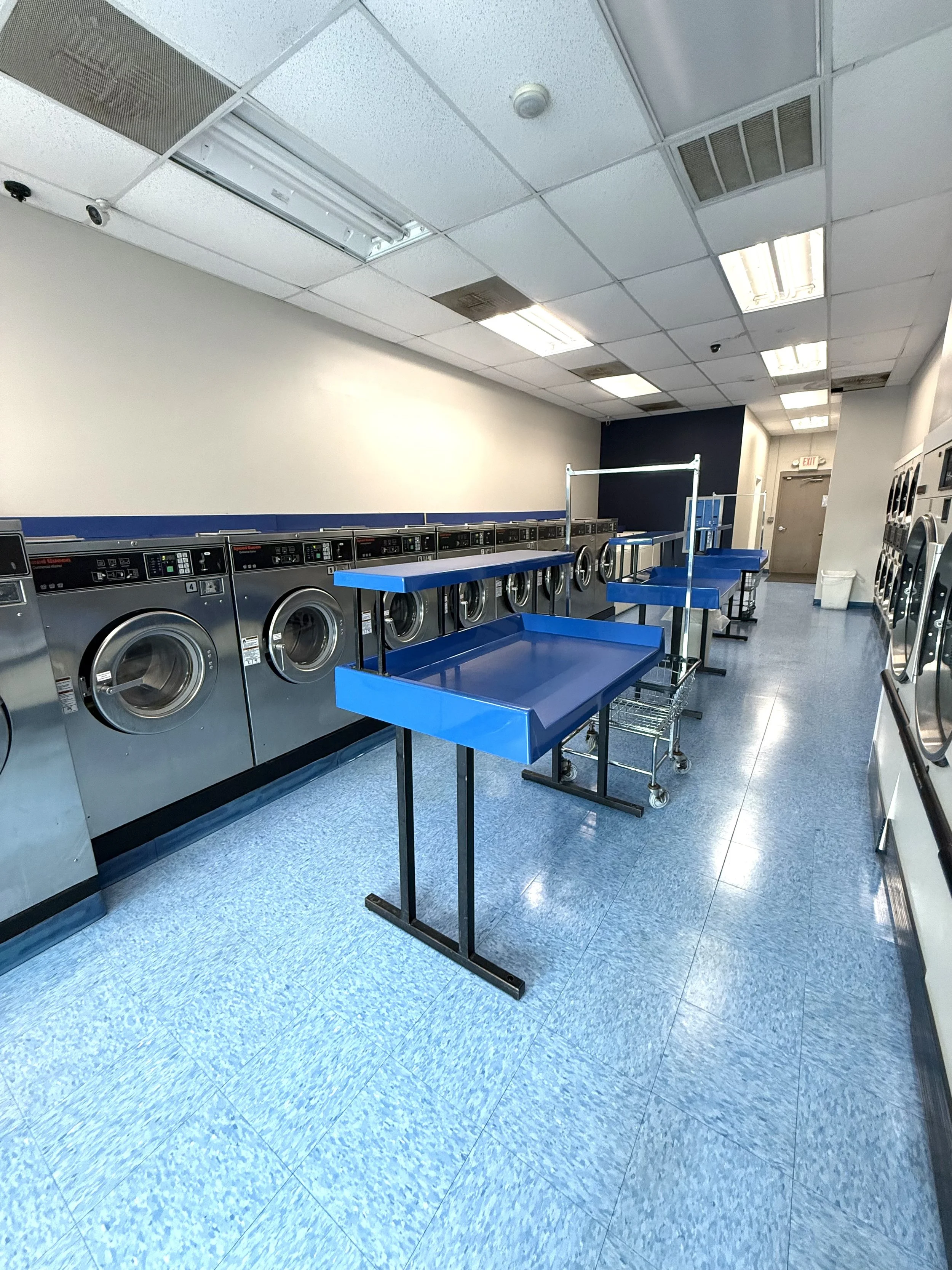 Laundry room with rows of washing machines, blue laundry carts, and a blue table, in a commercial laundry facility.