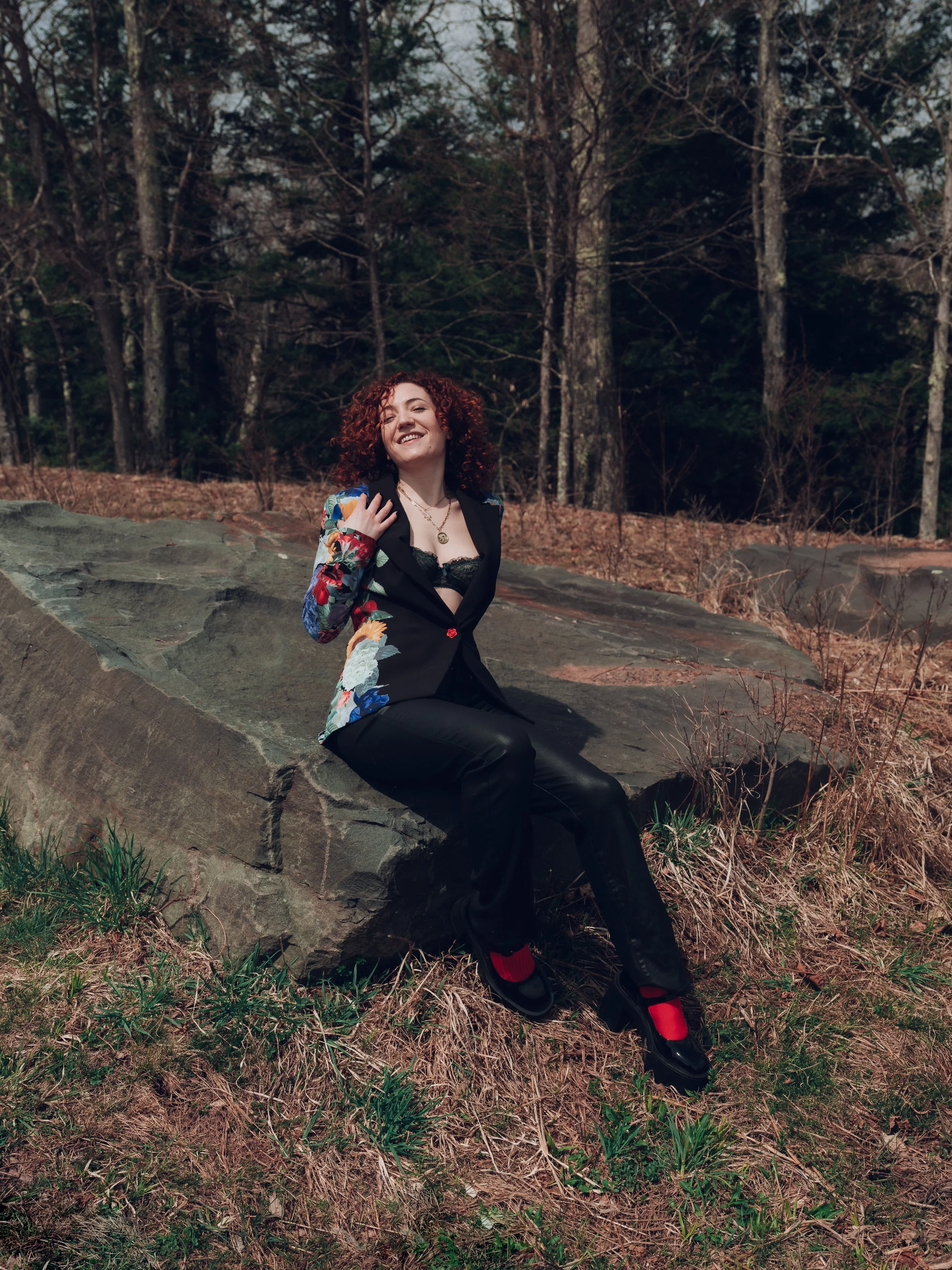 A woman with curly red hair is sitting on a large rock in a wooded area, smiling and looking happy.