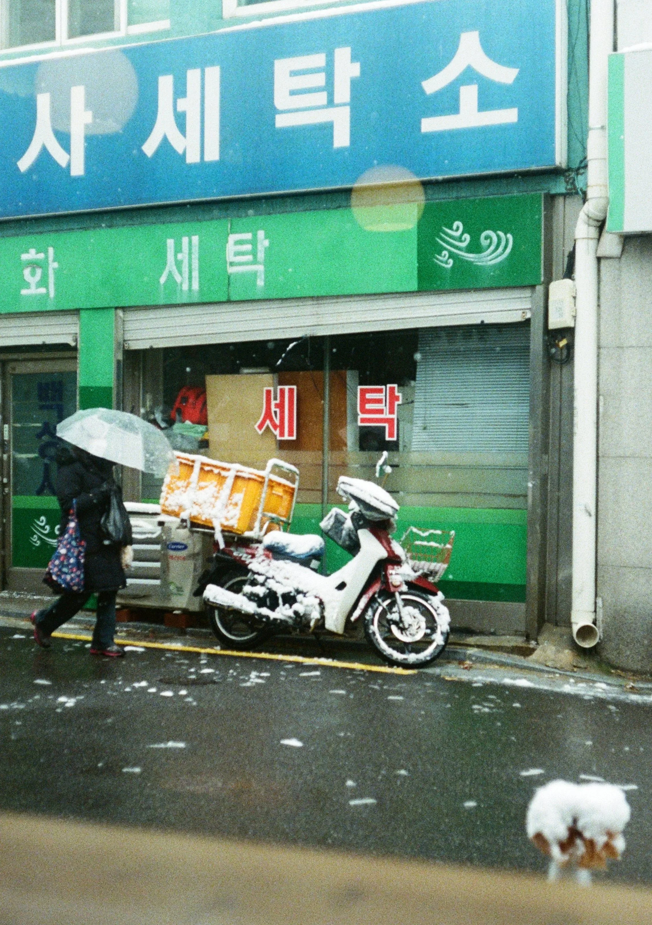 A woman walking past a snow-covered scooter and delivery cart parked outside a laundry shop with Korean signs on the storefront, and a person holding an umbrella nearby as snow falls on the street.