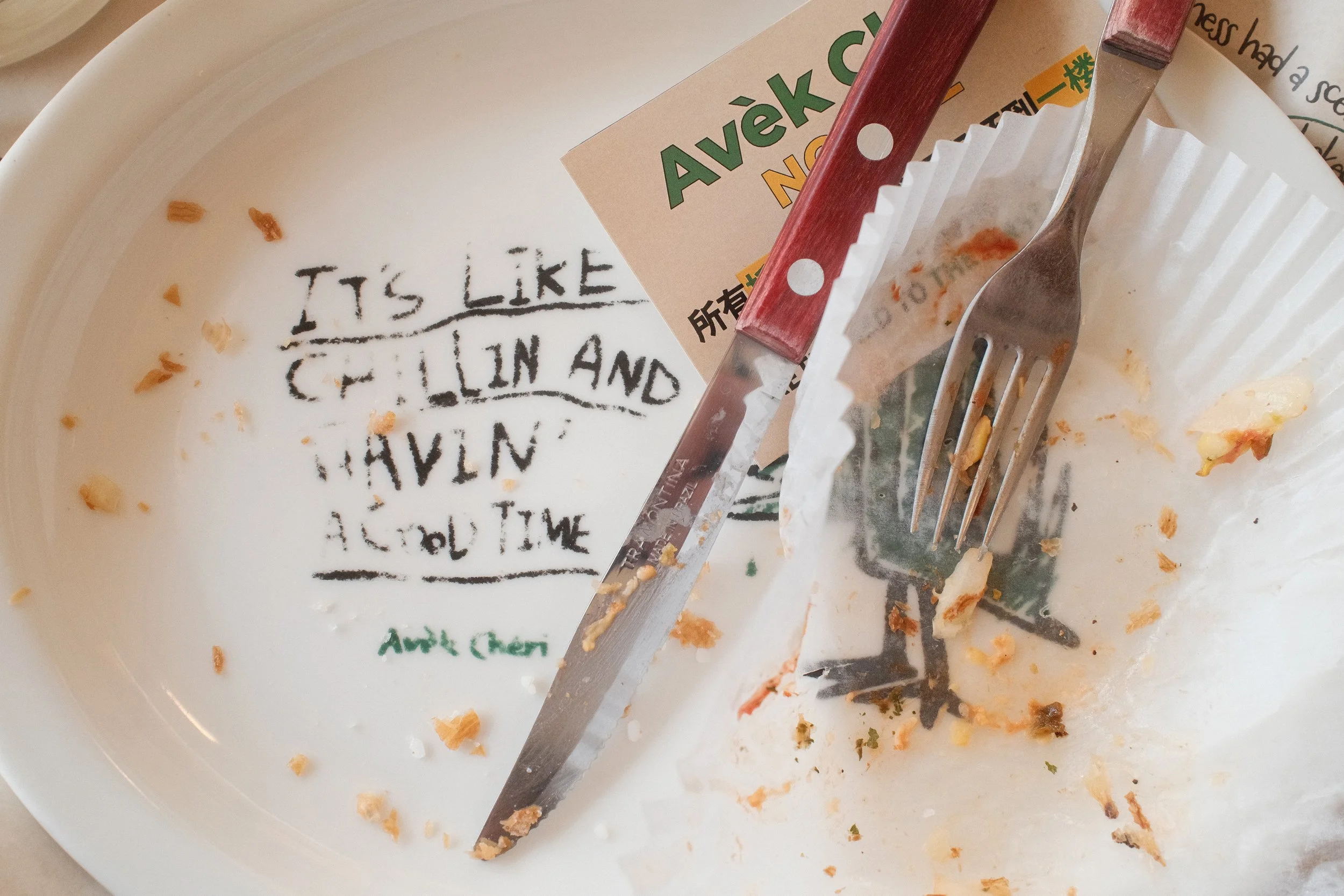 Empty white plate from Avek Cheri with the words 'It's like chillin and having a good time' handwritten in black ink. A fork and a knife rest on the plate, which also has a paper cupcake liner and some leftover food crumbs.