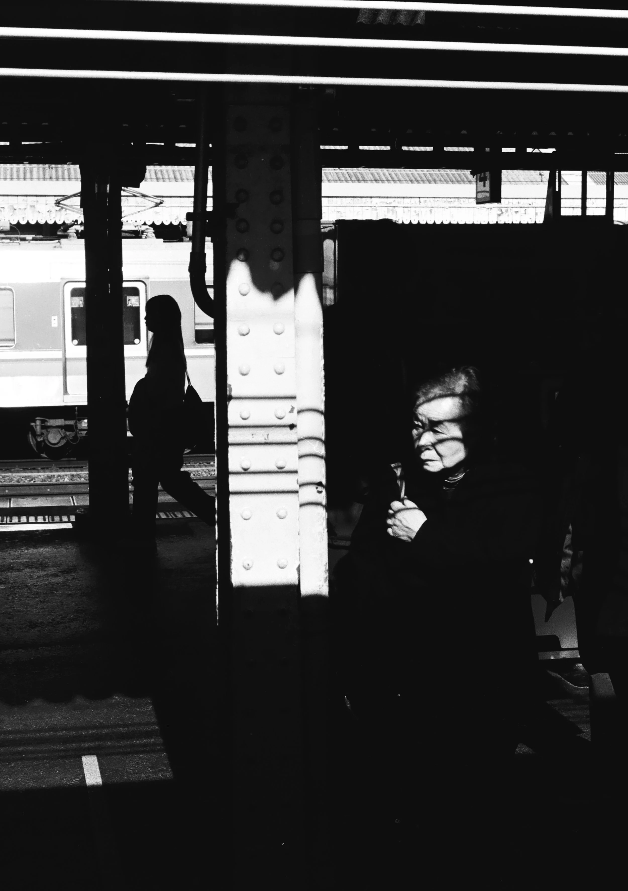 Black and white photo of a woman holding an umbrella on a train station platform, with a silhouette of a person standing in the background and a train passing by.