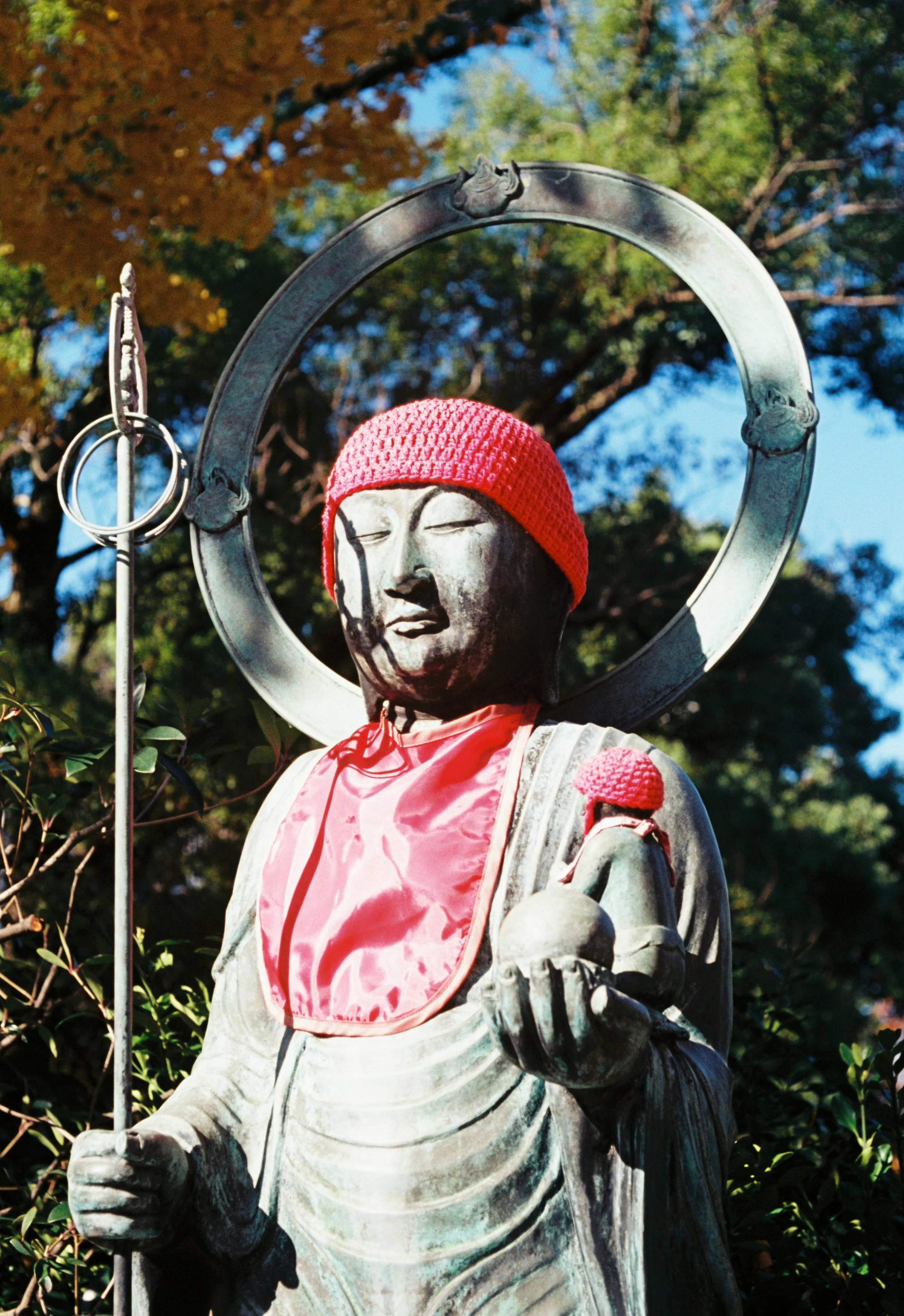A statue of a religious figure, possibly a Buddhist monk, wearing a red knit cap and a shiny, pink satin bib, holding a round object in one hand and a staff with hanging rings, surrounded by trees with blue sky in the background.