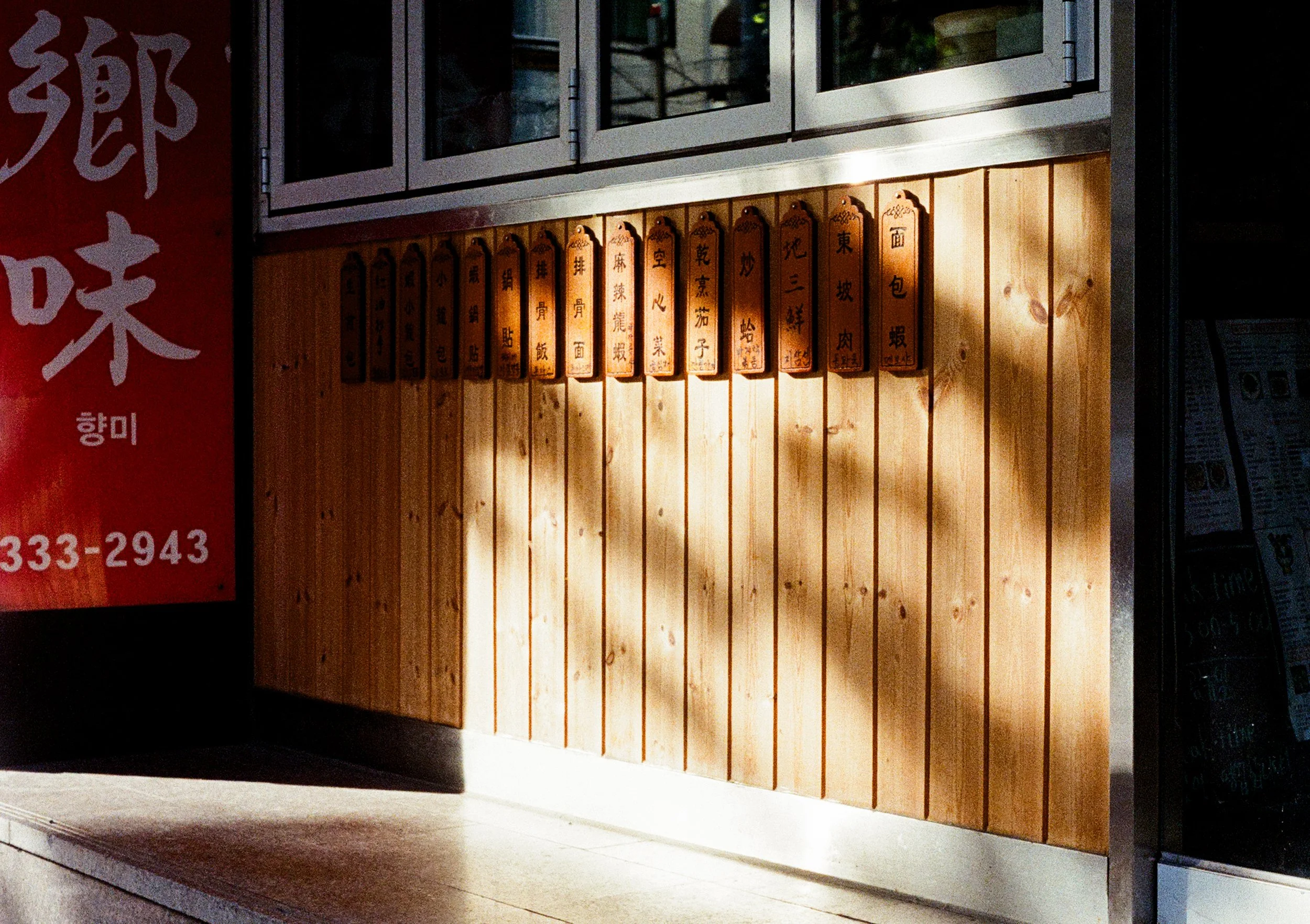 Wooden menu placards with Chinese dish names hang beneath a shuttered restaurant storefront, sunlight raking across the panels beside a red "鄉味" signboard. Shot on an Olympus Pen FT (38mm f1.8) with Kodak Ultramax 400, processed by Yeonnam Film.