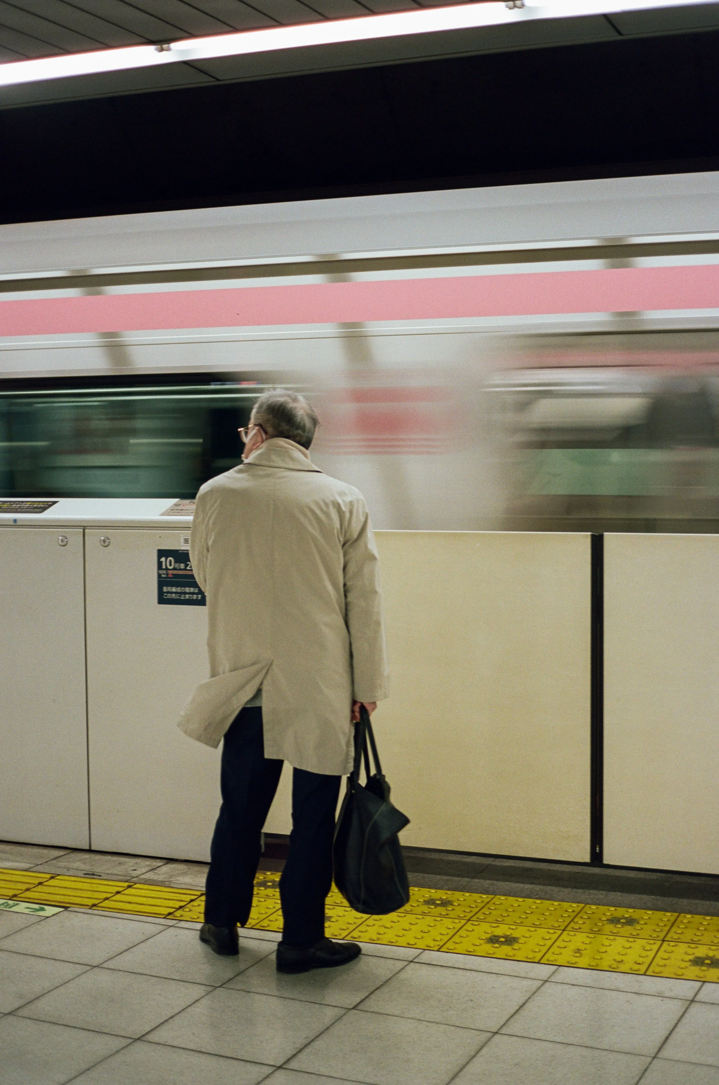 A man wearing a beige trench coat, dark pants, and glasses standing on a subway platform, holding a black bag, as a train speeds by in the background.