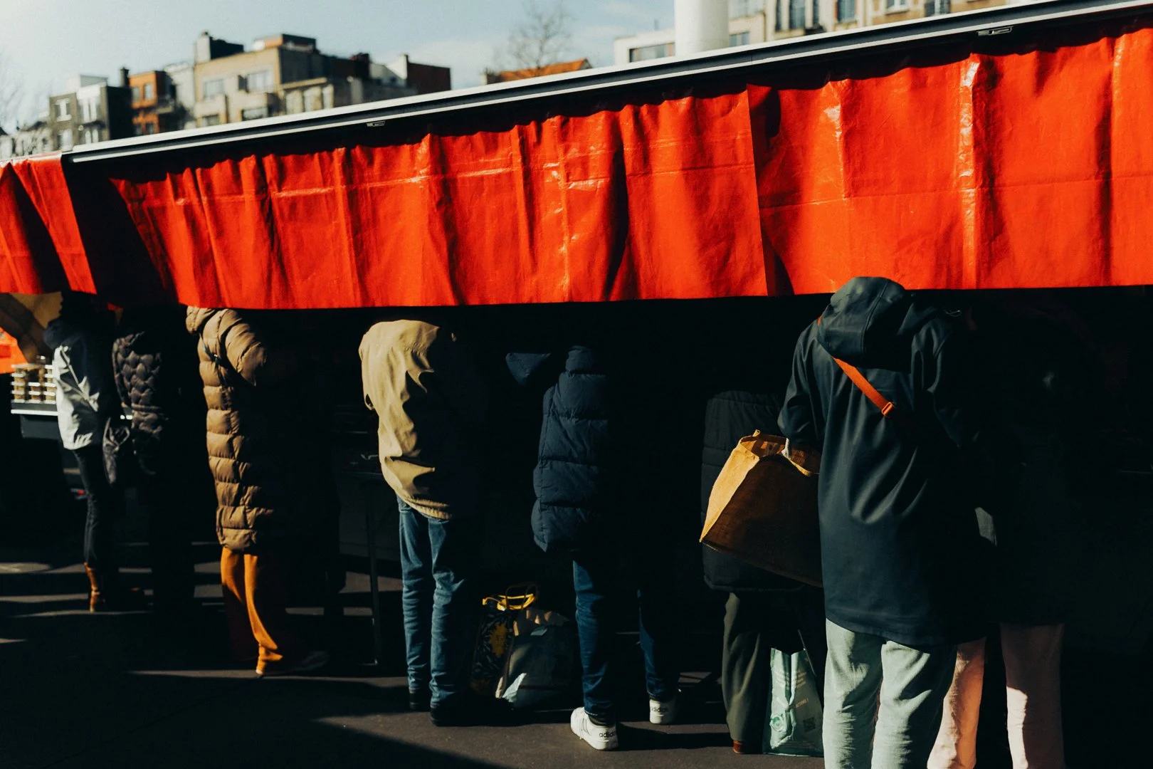 People standing in line at an outdoor market stall with a red canopy, in an urban setting with apartment buildings in the background.