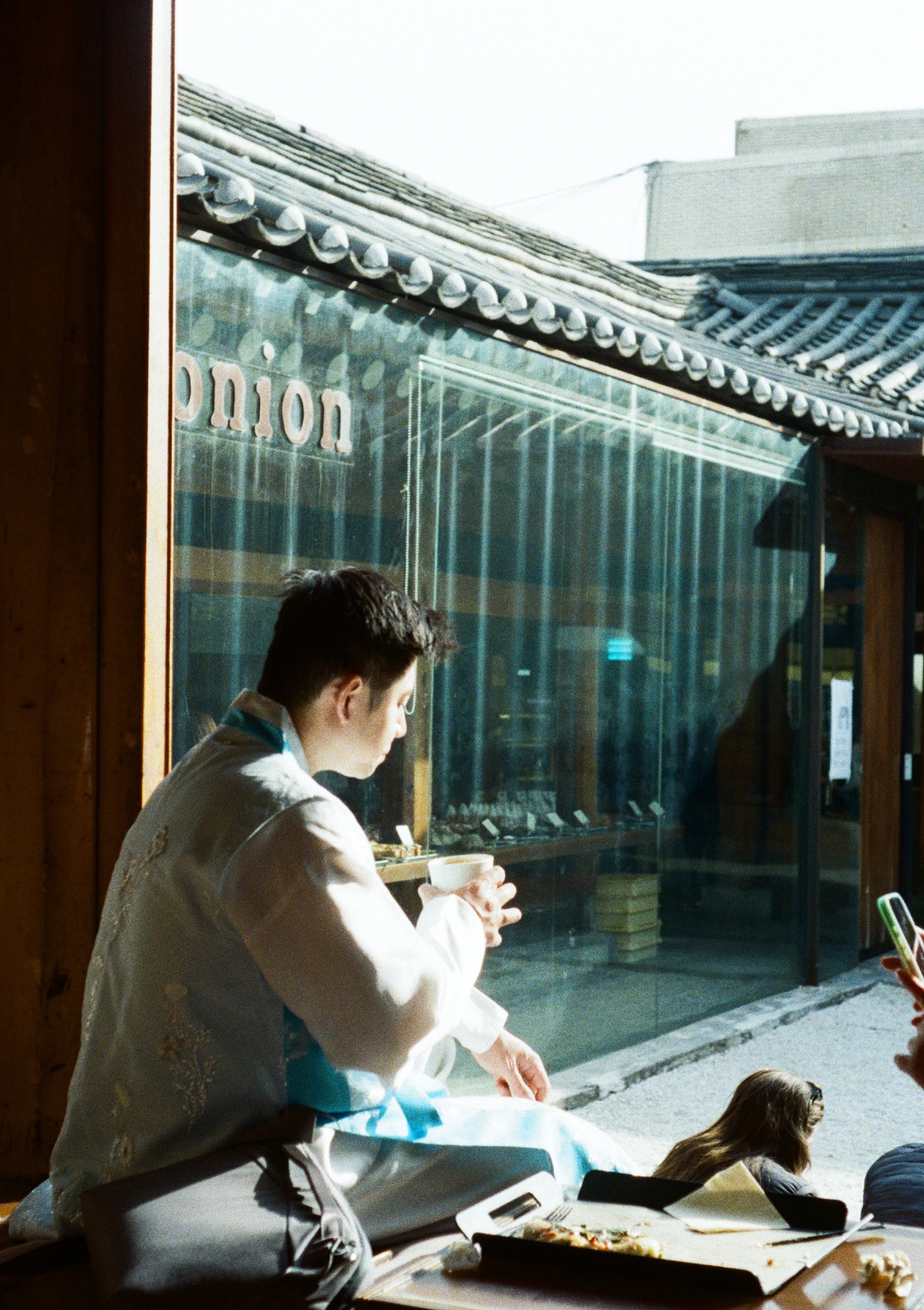 A person dressed in traditional Korean hanbok sitting by a window, holding a cup, with food on the table. Outside the window are buildings with tiled roofs.