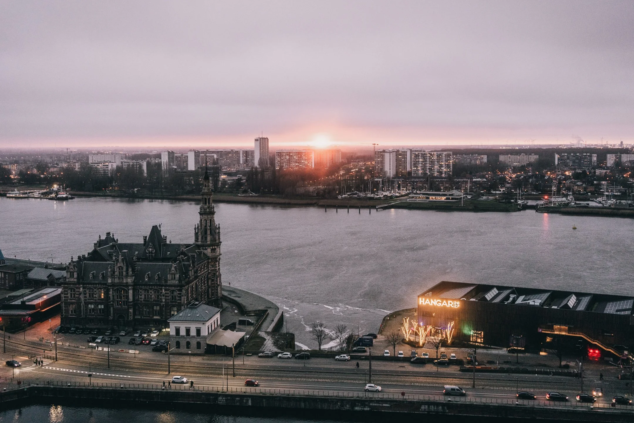 Cityscape at sunset featuring a river, historic building, modern buildings, and an illuminated sign that reads 'HANGAR126'.