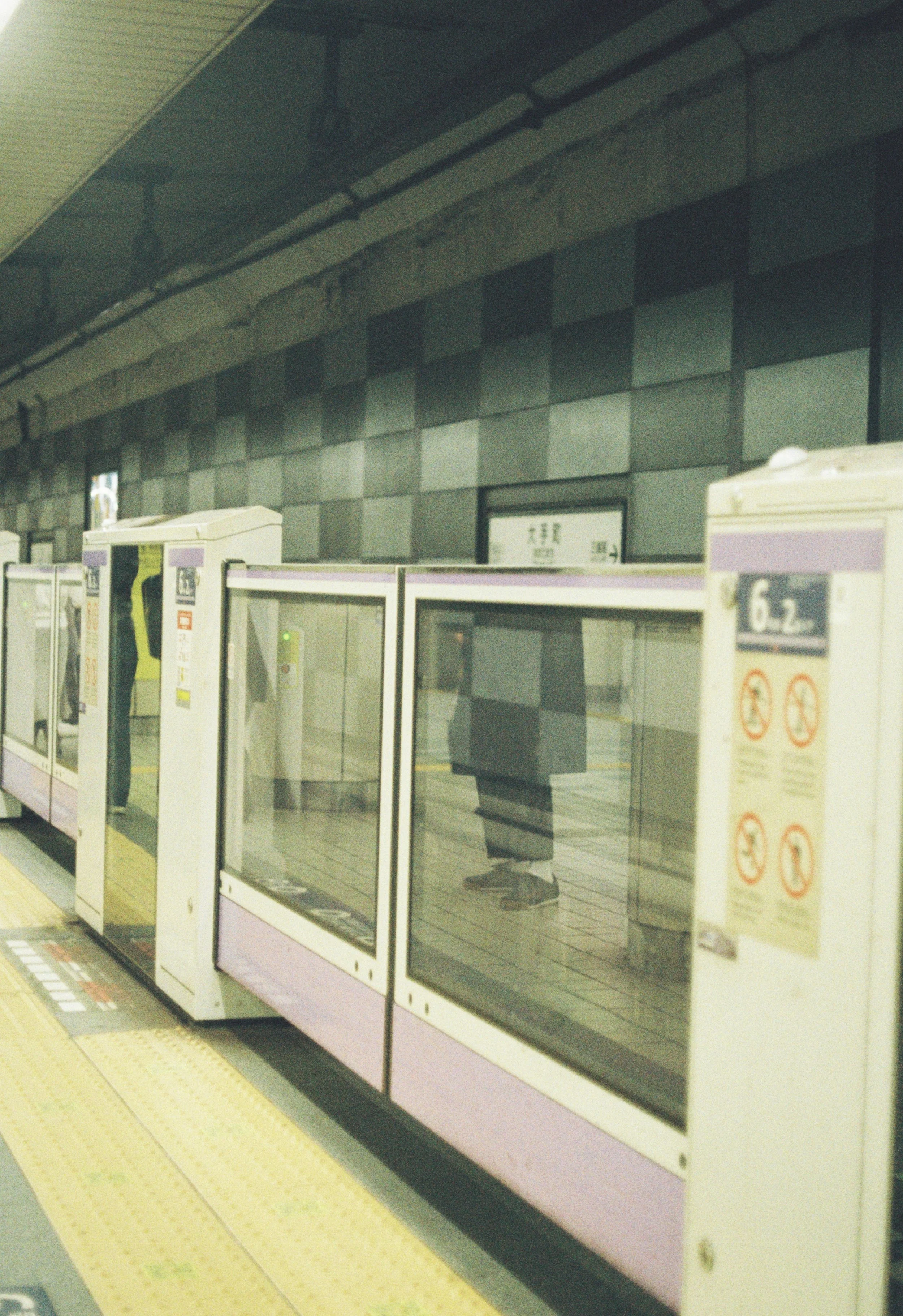 View of a subway station platform with glass barriers and a person standing behind them, visible through the glass, wearing sneakers and pants. The platform has yellow tactile paving for the visually impaired.