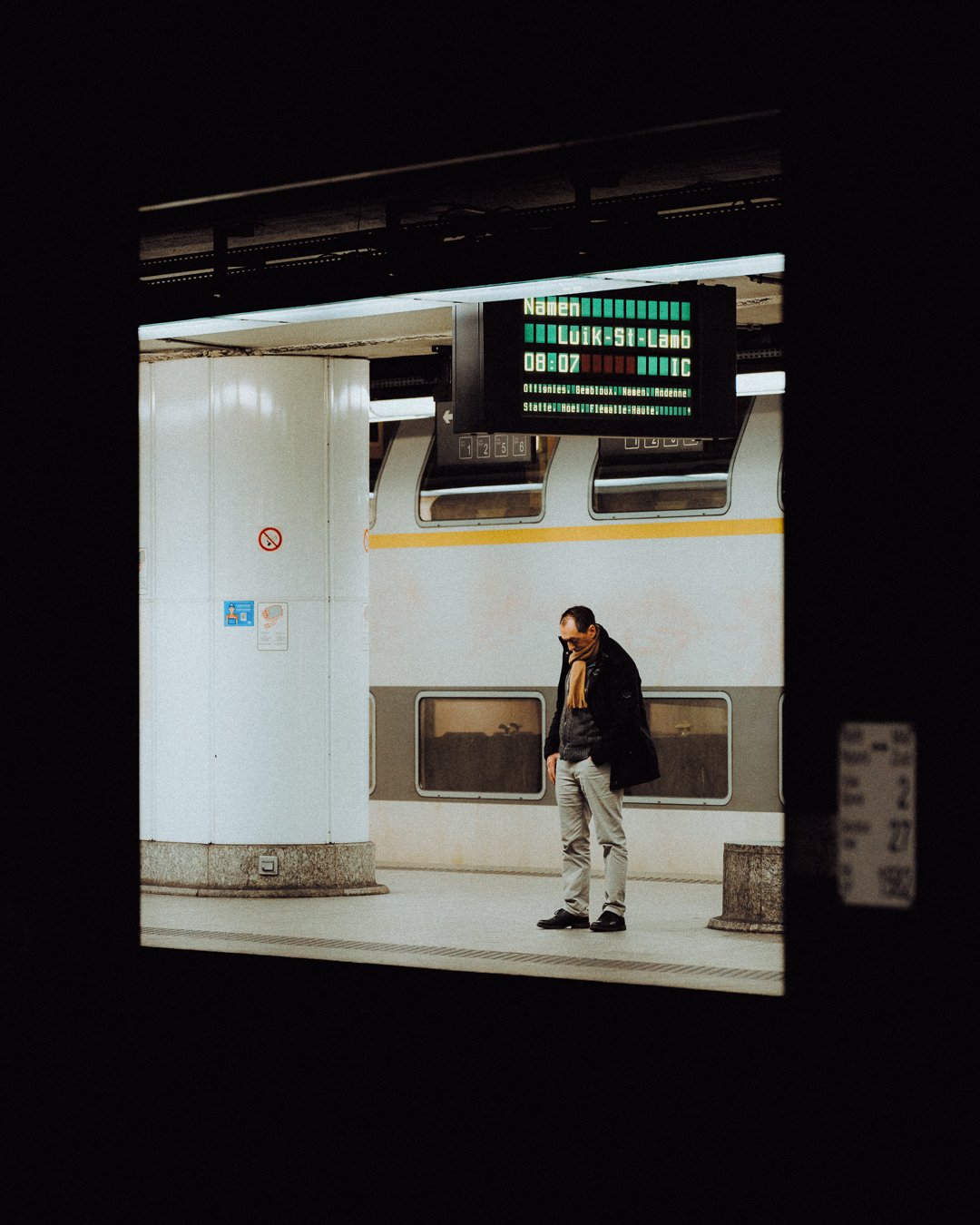 A man standing on a train platform talking on his phone, with train cars behind him and an electronic timetable above displaying station information.