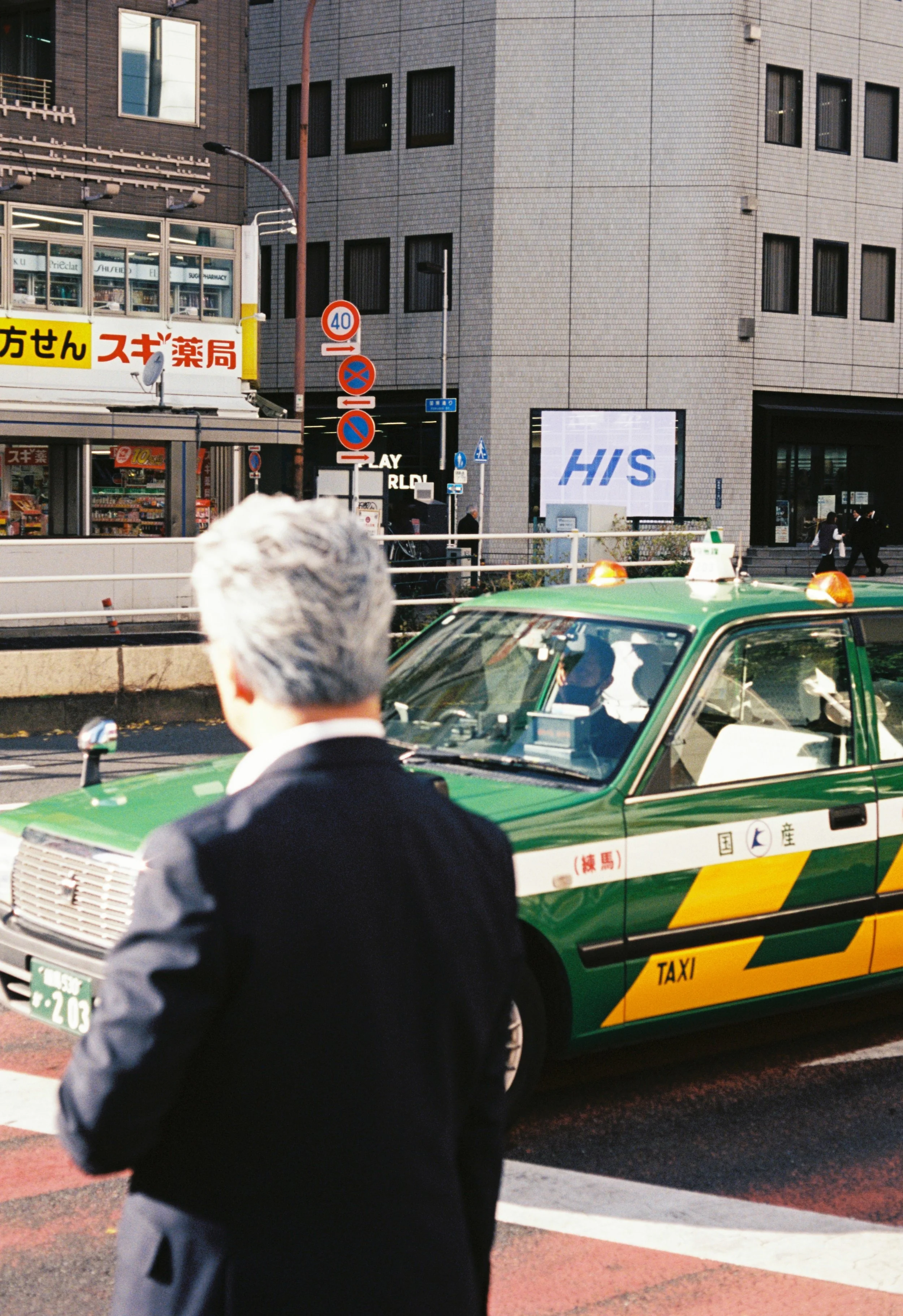 A man with gray hair, dressed in a dark suit, standing in front of a green taxi cab on a city street. There are storefronts, street signs, and a tall building in the background.