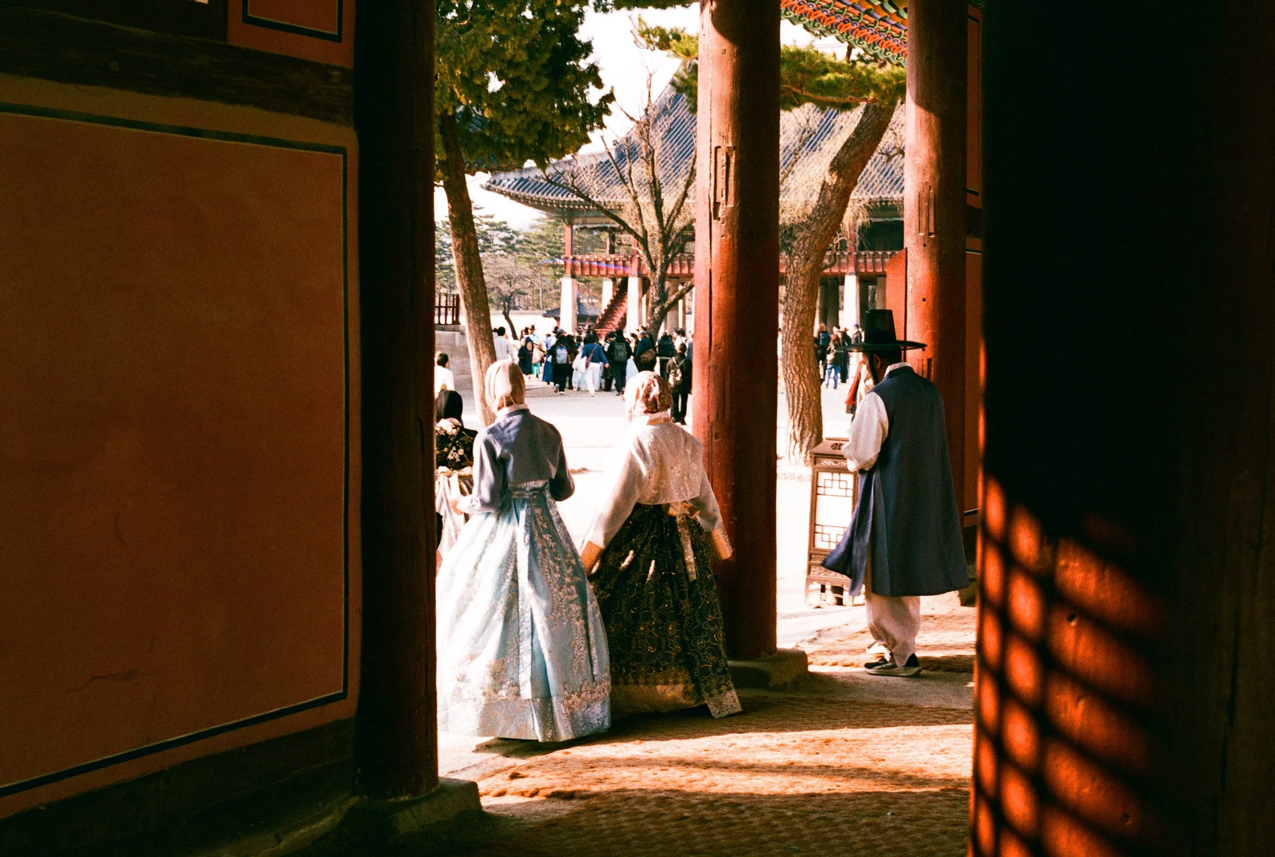 2 Women and a man stepping into light from a shadowy location while dressed in traditional Korean Hanbok at Gyeongbokgung Palace. Taken on Lucky Color 200 Film with Contax G1.