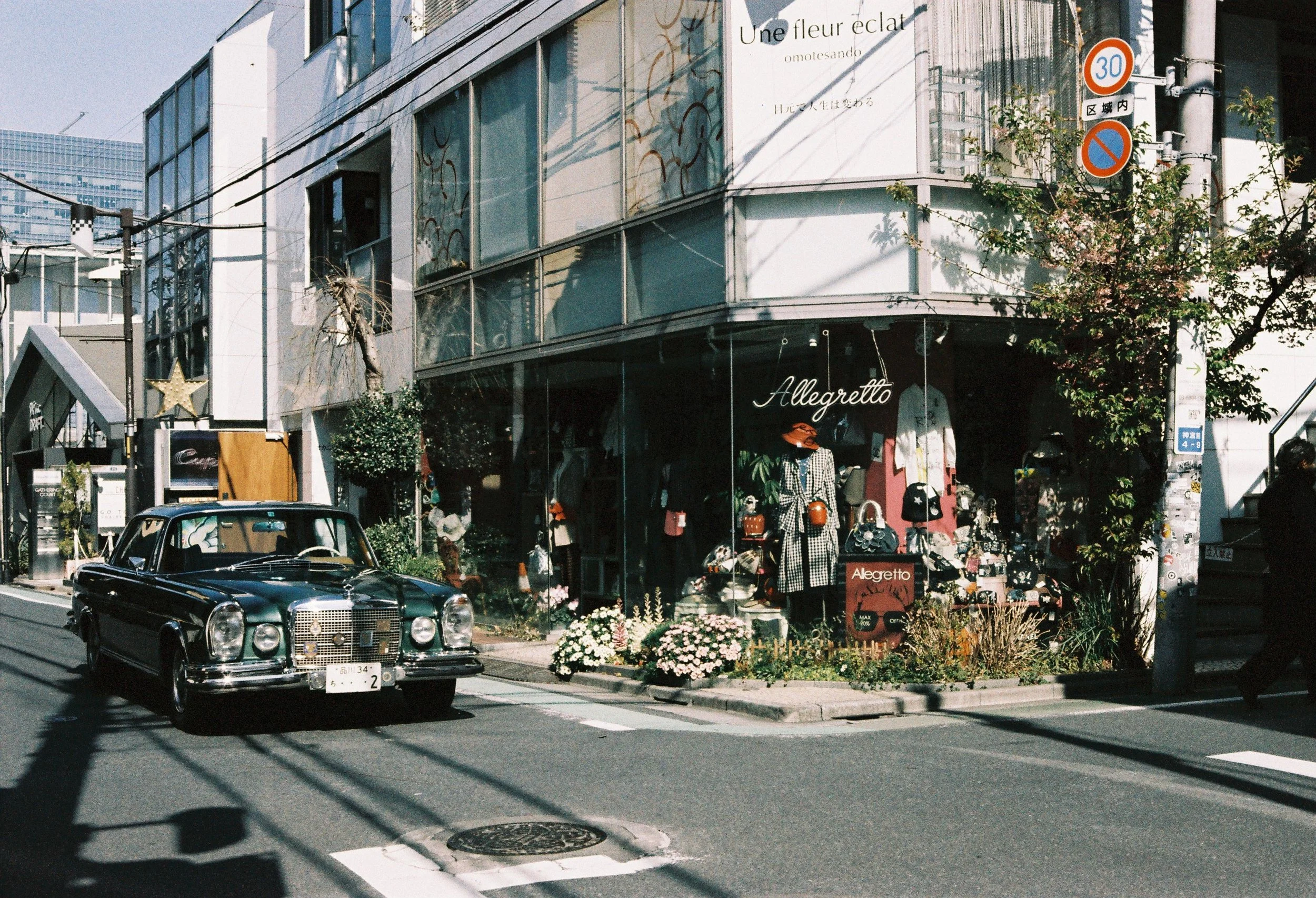 A vintage black car parked on a city street in front of a boutique clothing store named 'Allegretto', with mannequins dressed in fashionable clothing and accessories displayed in the window. The store is decorated with plants and flowers, and a sign 