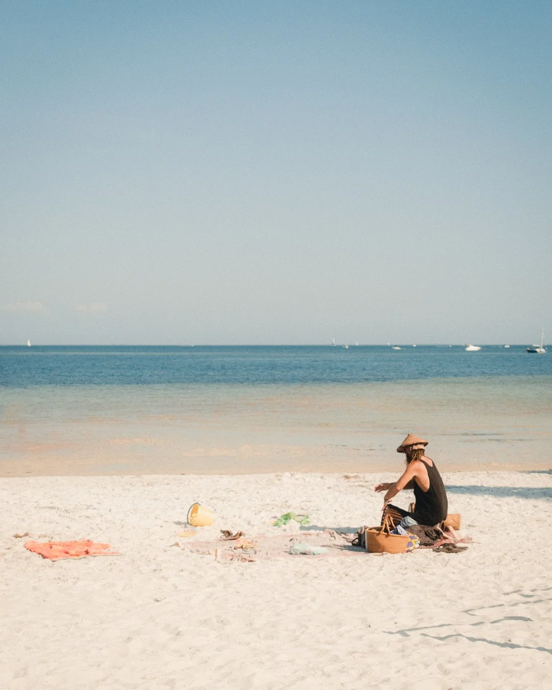 A woman sitting on the sandy beach with a straw hat, surrounded by beach items, with ocean and sailboats in the background.