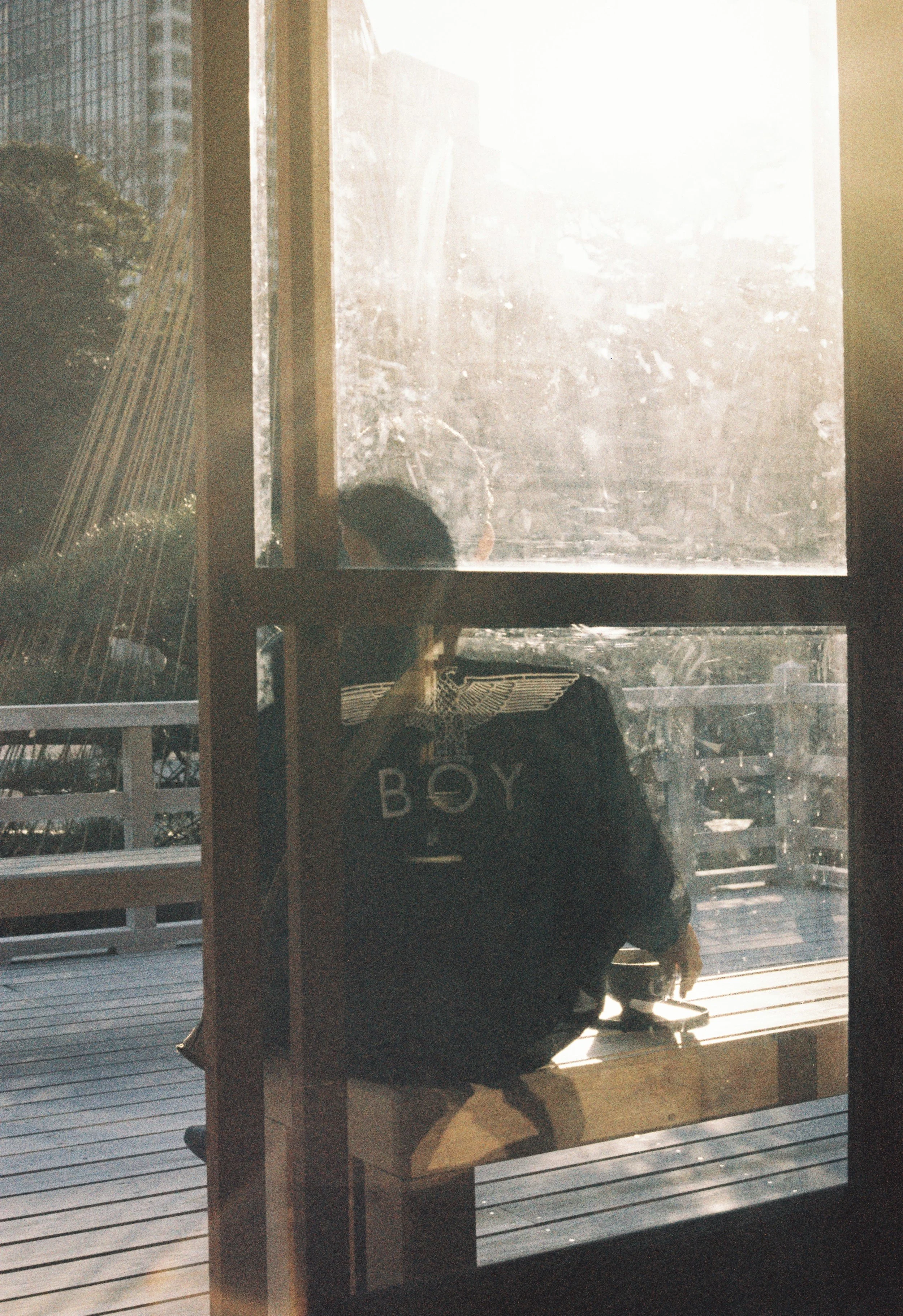 A person sitting on a wooden bench inside a room, viewed through a glass door, with sunlight shining and a cityscape background.
