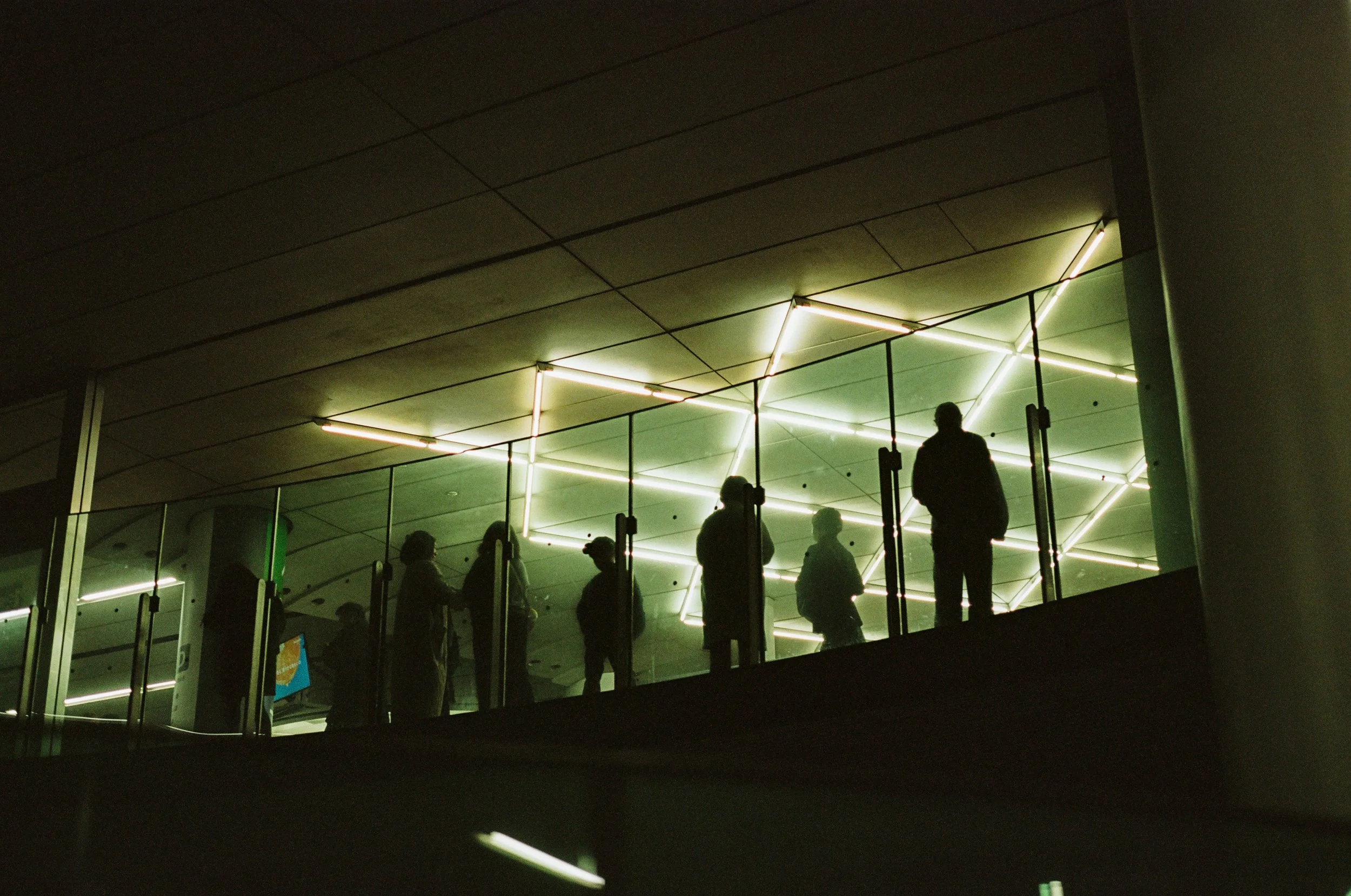 Silhouettes of people standing on a balcony with modern lighting fixtures on the ceiling, inside a building.