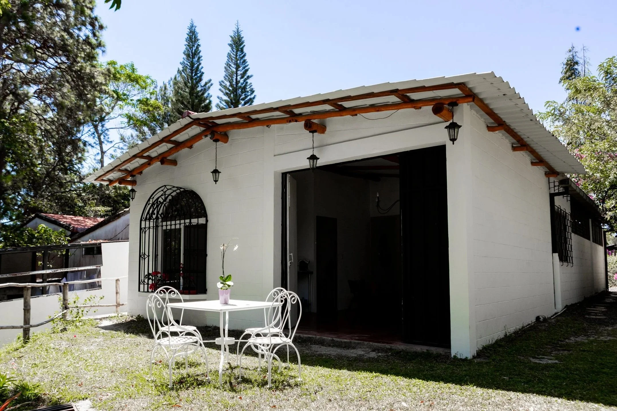 Área de terraza bajo un árbol grande, con muebles metálicos y una mesa con mantel de cuadros rojos. En el fondo, una casa de ladrillos con ventanas y una cerca de metal.