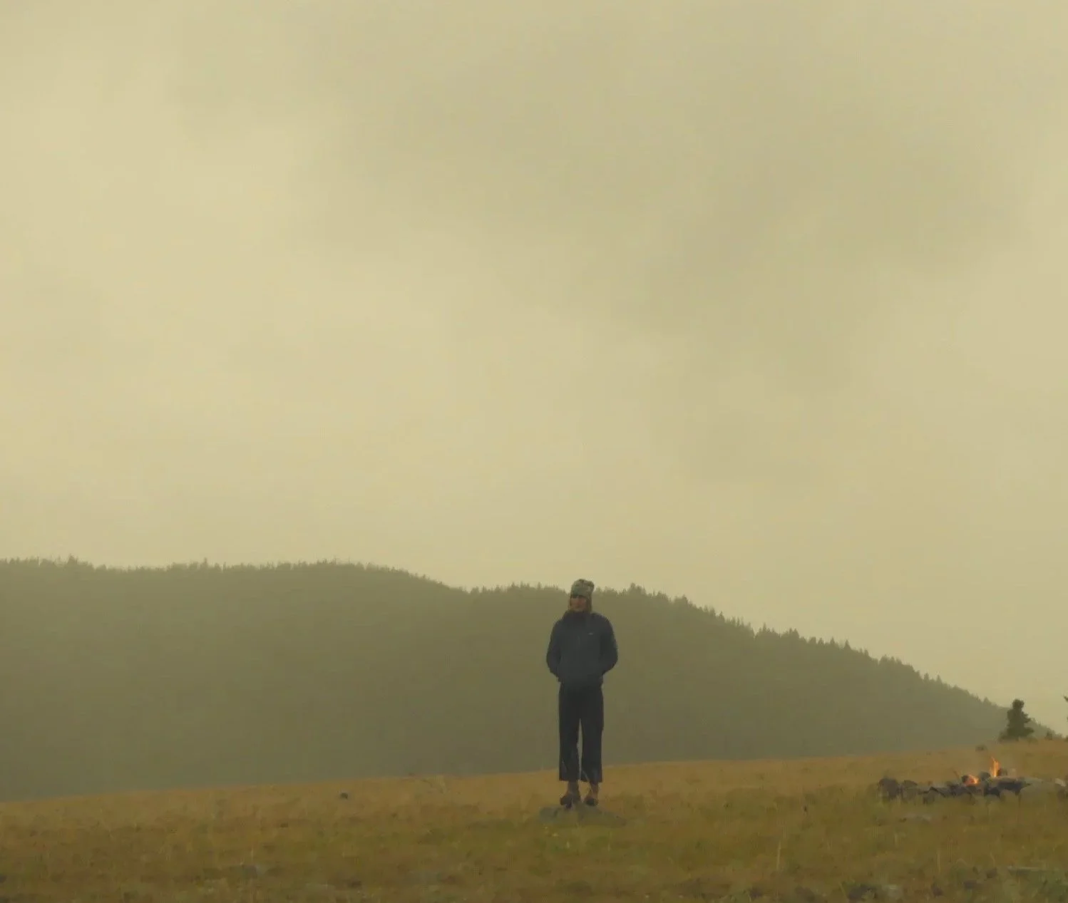 Person standing on a grassy field near a small campfire, with a backdrop of forested hills and a hazy sky.