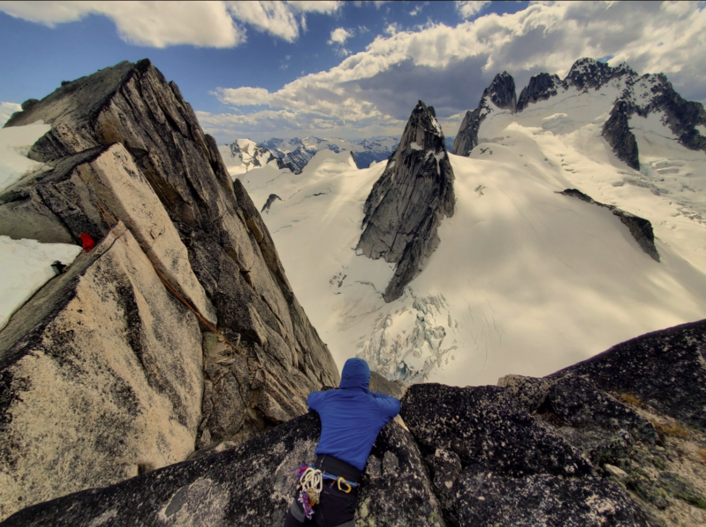 A climber in a blue jacket and harness is resting on a rocky ledge in a snowy mountain landscape, with towering snow-capped peaks and a cloudy sky in the background.