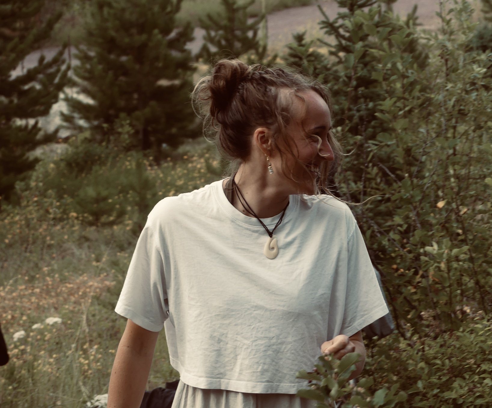 A woman with curly hair in a bun, wearing a white t-shirt, smiling and outdoors amidst trees and greenery.