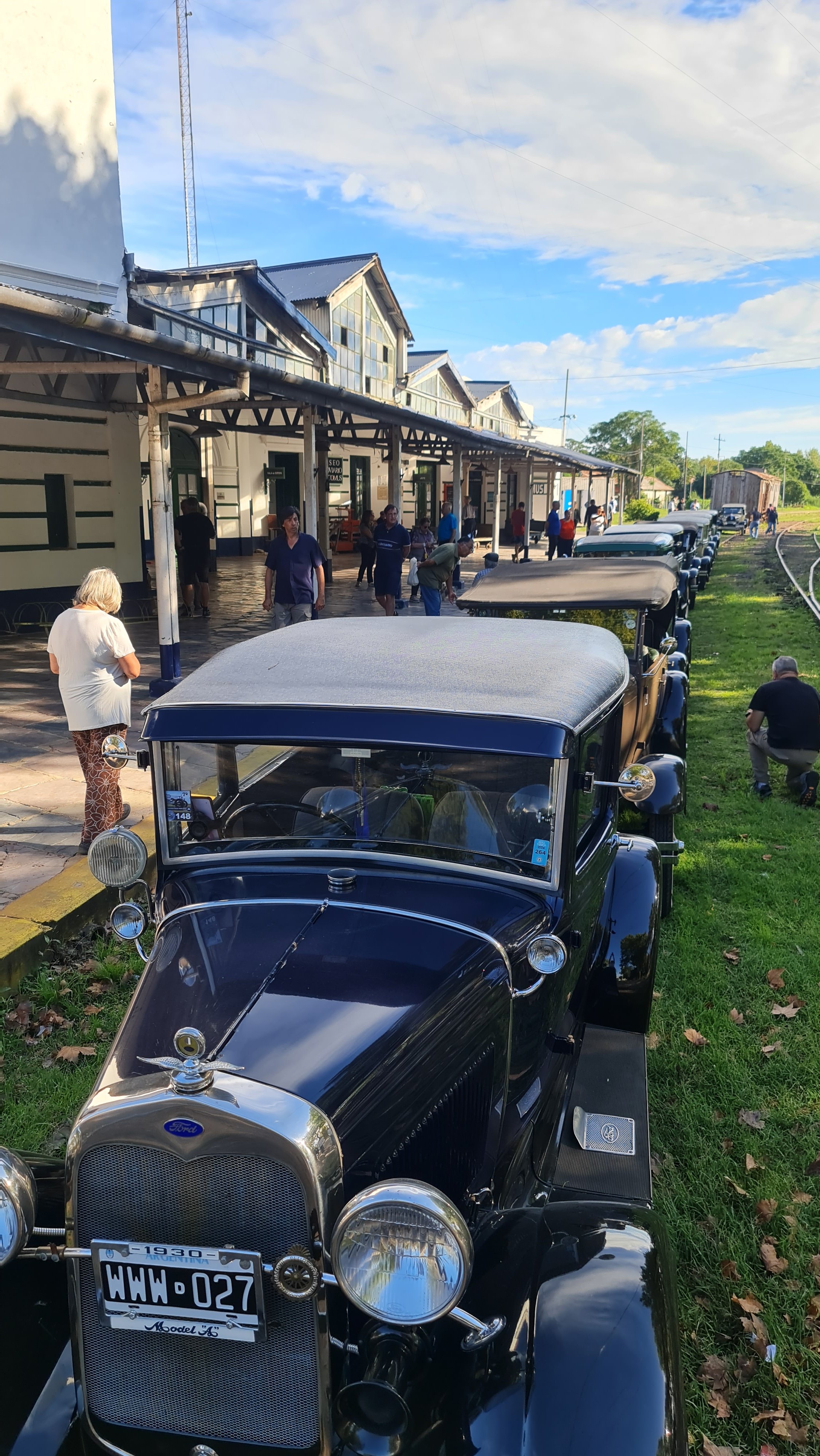 Antiguos autos en una fila en una estación de trenes, con un edificio de estilo clásico y personas caminando en la plataforma, cielo despejado.