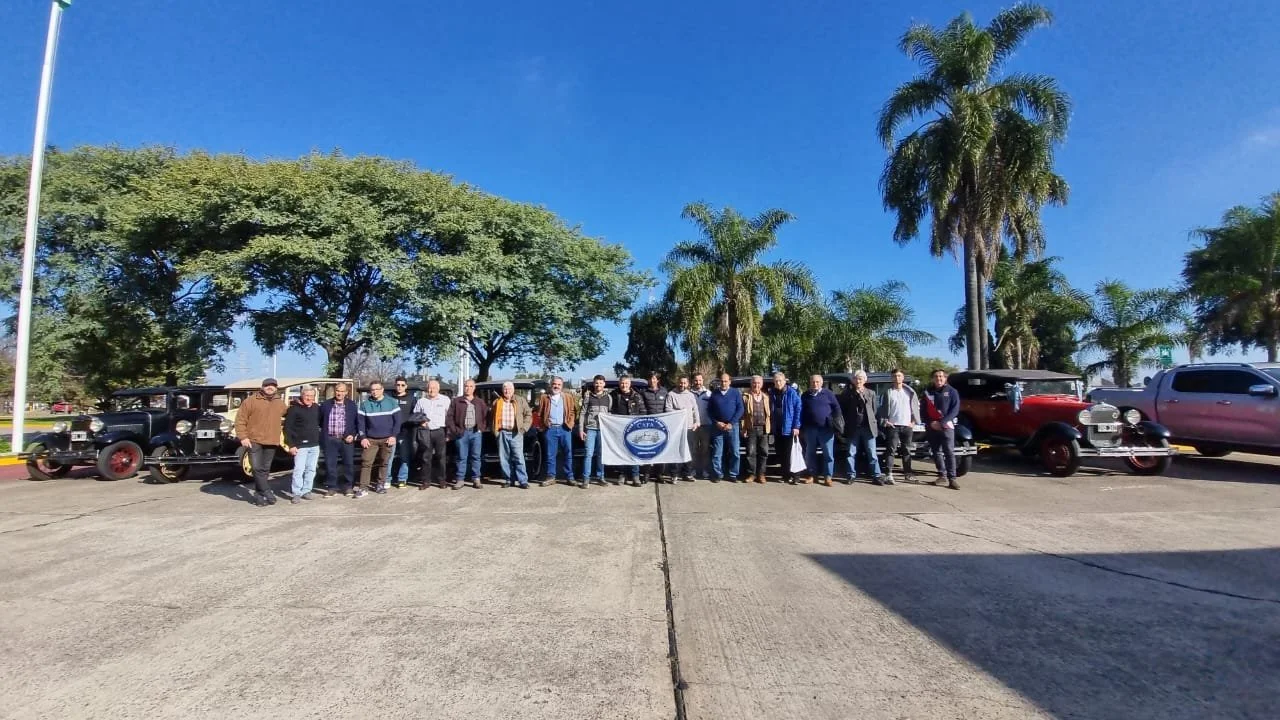 Grupo de personas frente a autos antiguos, en un entorno con árboles y palmeras, bajo un cielo despejado.