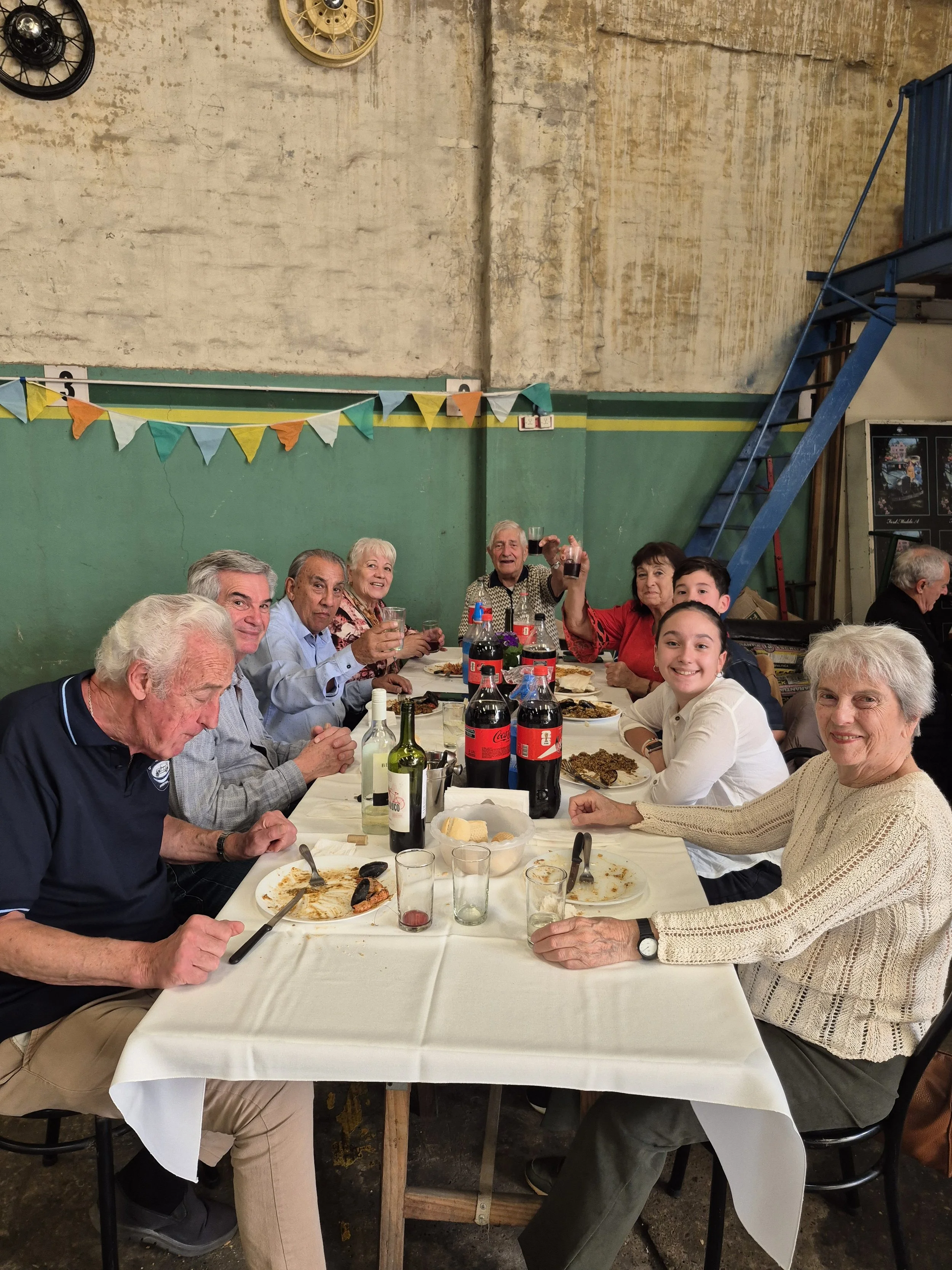 Grupo de personas reunidas en una mesa comiendo y brindando en un ambiente interior, con decoraciones coloridas y paredes de color verde y beige.