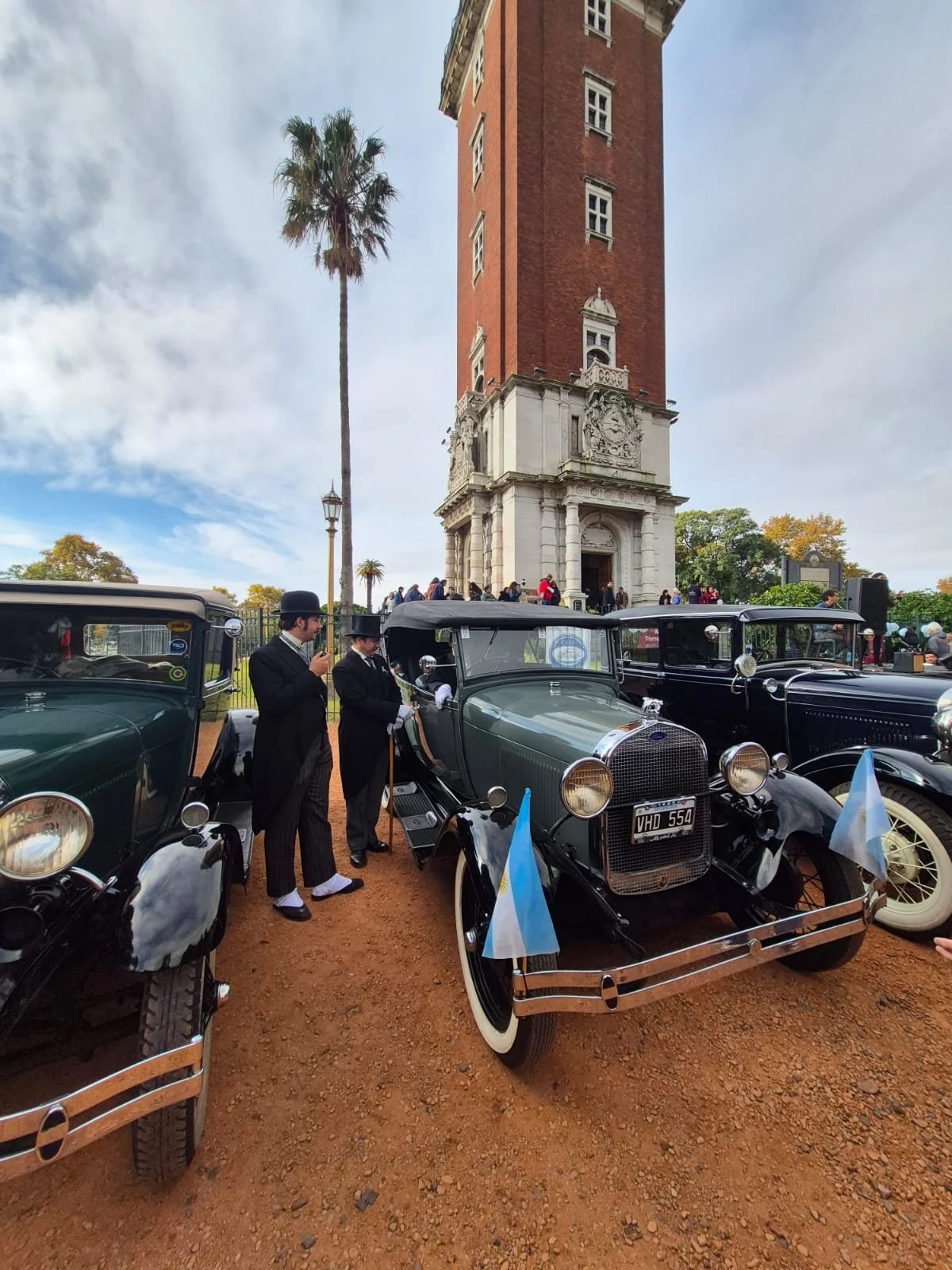 Personas vestidas con ropa de época junto a autos antiguos frente a una torre de estilo colonial en un día soleado con algunos árboles y un grupo de personas en el fondo.