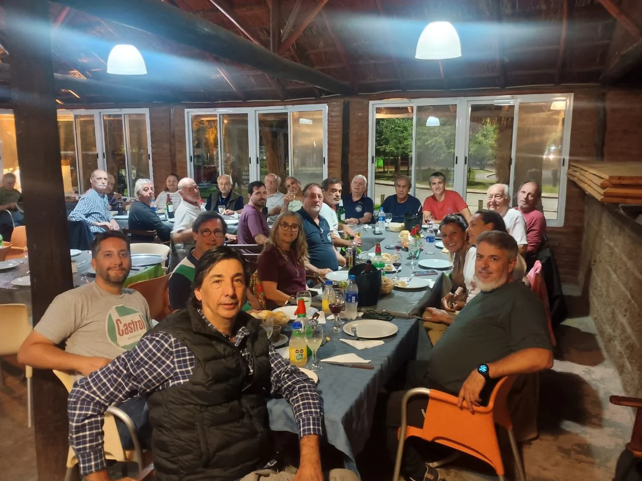 Grupo de personas en una reunión o comida en un comedor con paredes de madera y ventanas grandes.