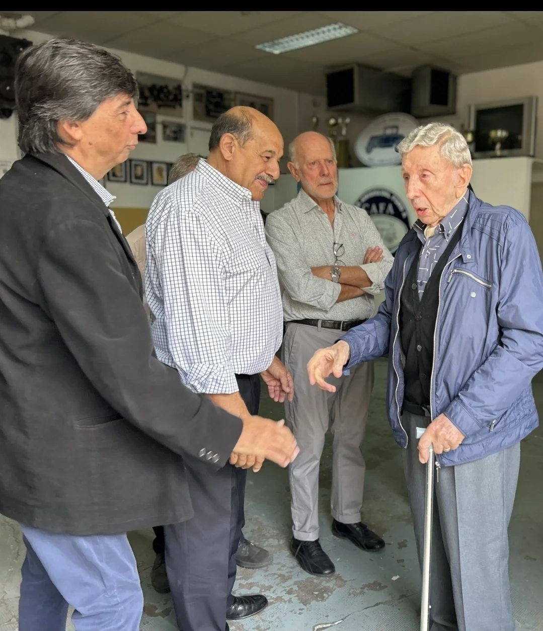 Grupo de cinco hombres adultos conversando en un entorno interior, con uno de ellos usando un bastón.