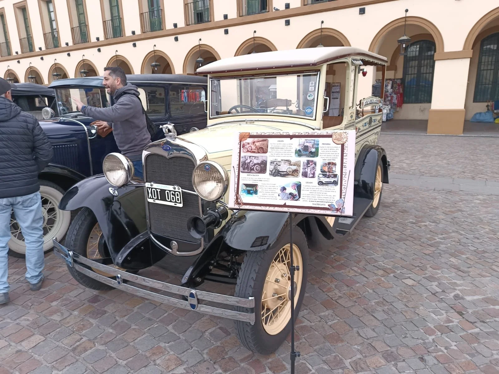 Coche antiguo de exhibición en una plaza, con un cartel informativo y varios espectadores conversando.